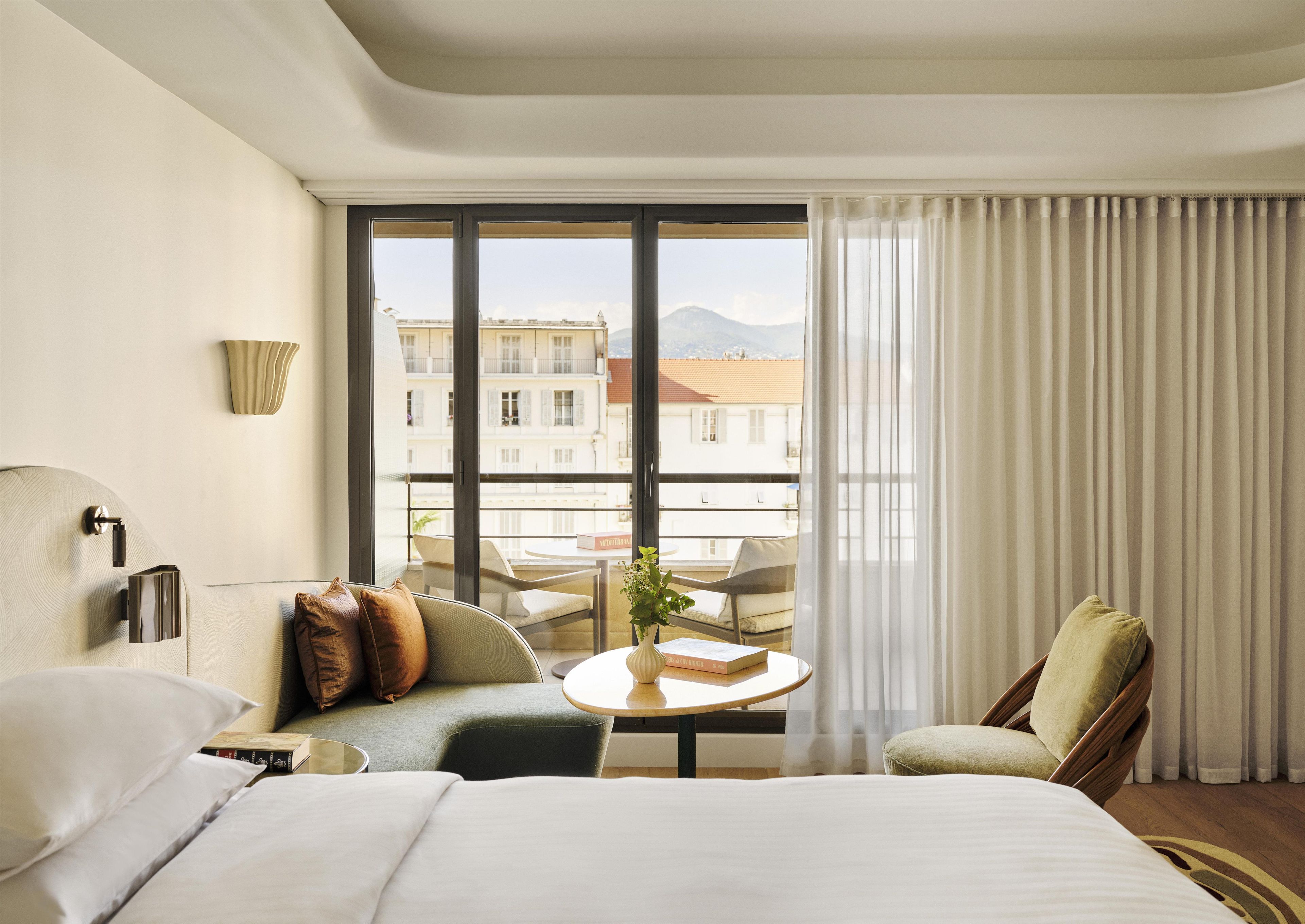 Ett hotellrum med en säng, en soffa, en stol, ett litet bord med en bok och utsikt över byggnader genom stora fönster.