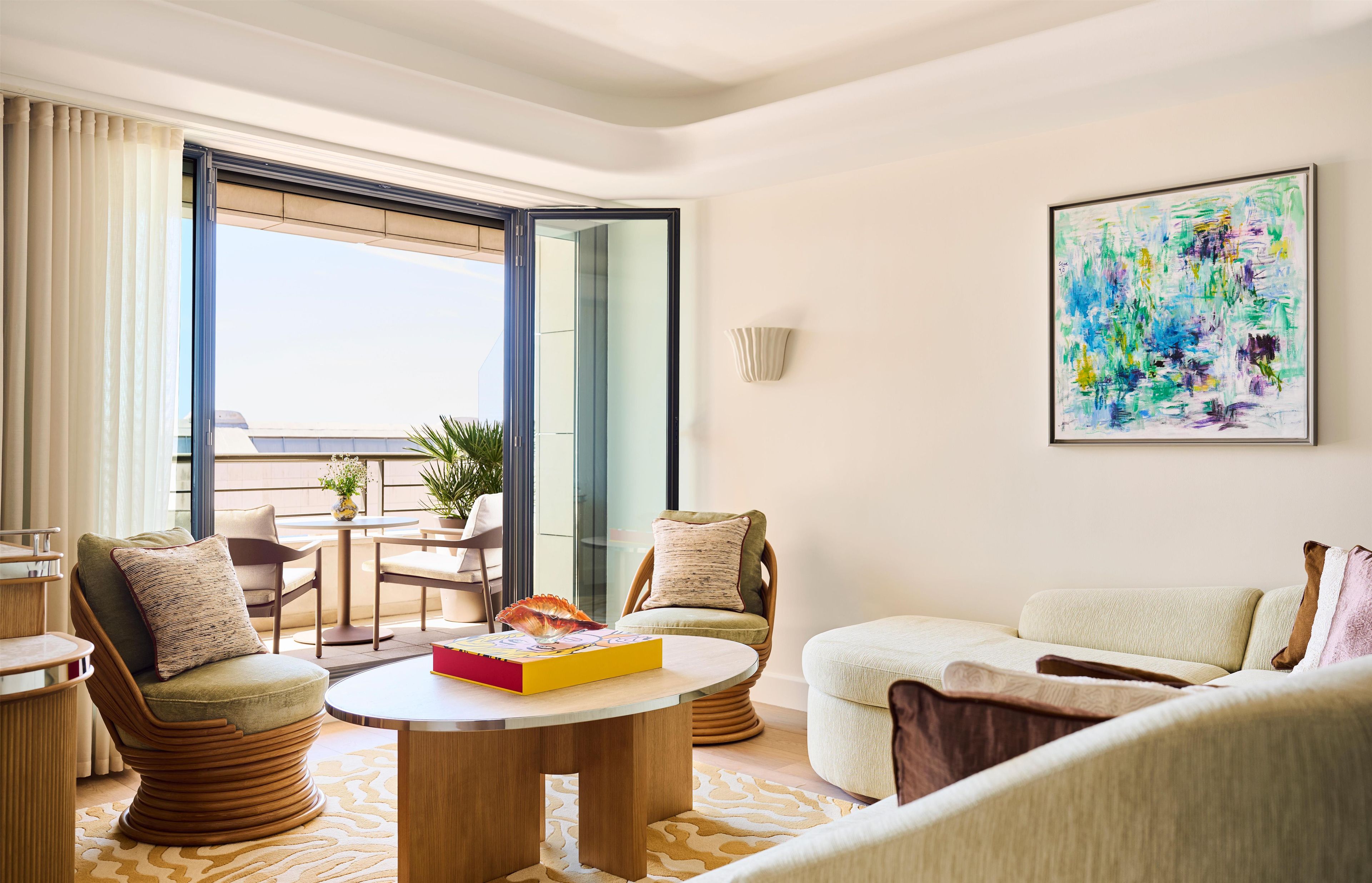 A modern living room with a round coffee table, two armchairs, a sofa, a dining table with chairs, a large window, and a colorful abstract painting on the wall.