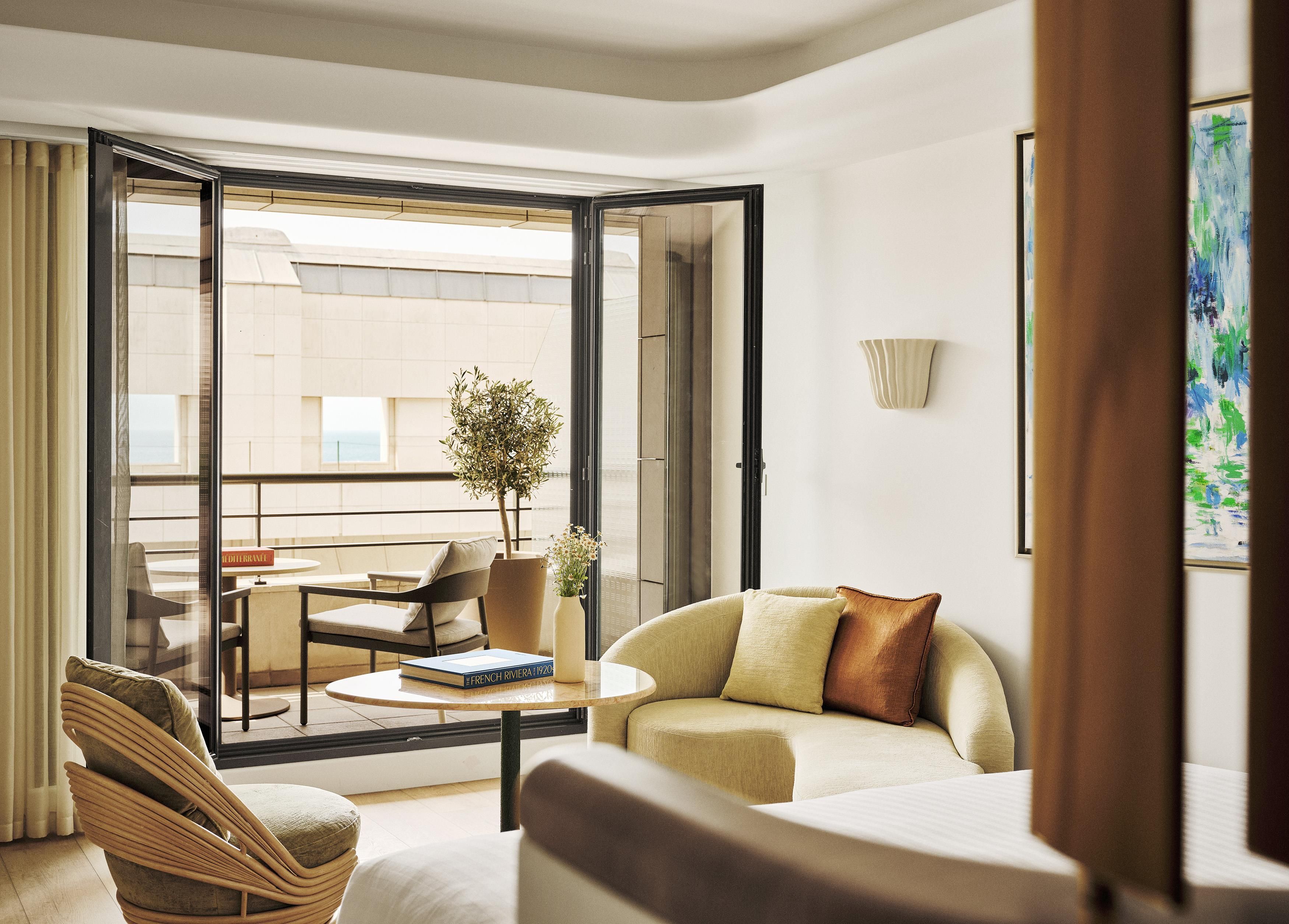 A modern hotel room with a balcony, a sofa, a chair, a small table, and a potted plant.
