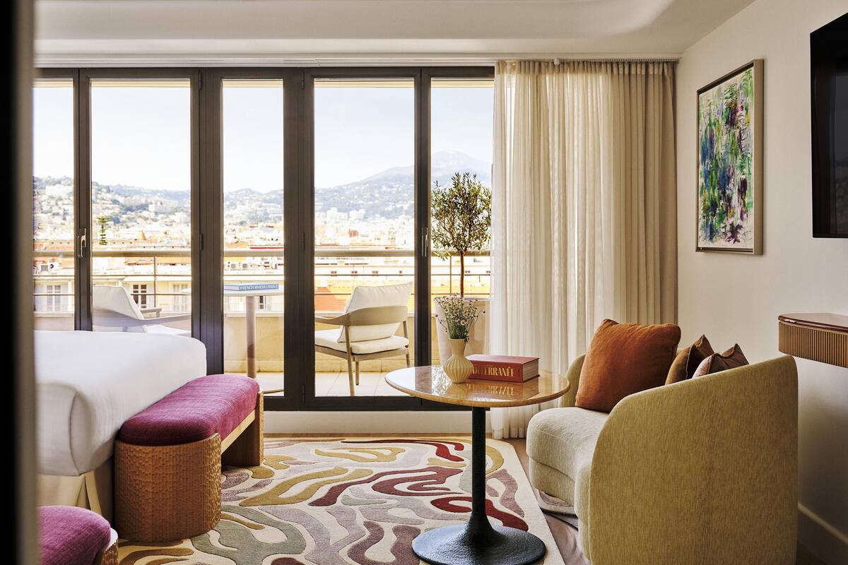A hotel room with a balcony, a bed, a sofa, a small table, and a painting on the wall.