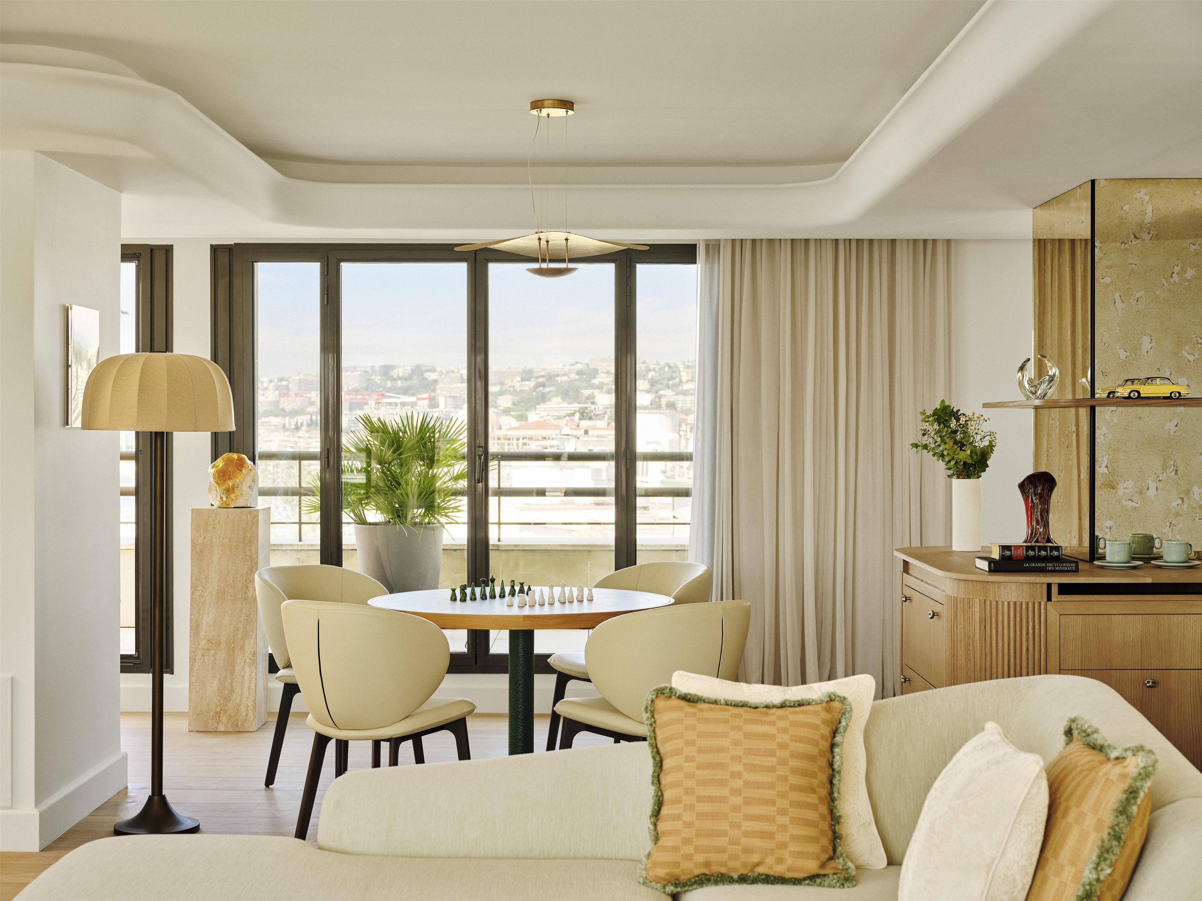 Un salón moderno con sofá, mesa de comedor, sillas, lámpara y vista a la ciudad a través de amplias ventanas.