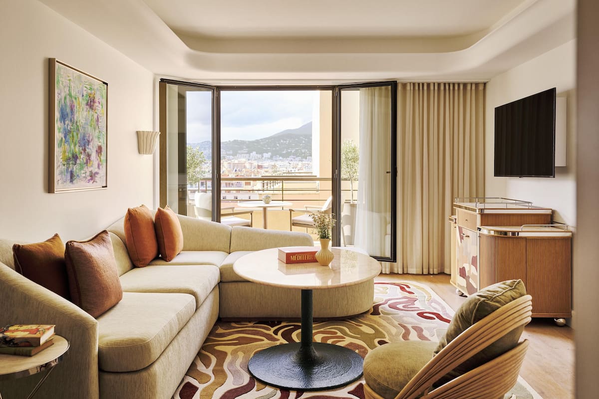 A modern hotel room with a sofa, a round table, a TV, and a balcony with a city view.