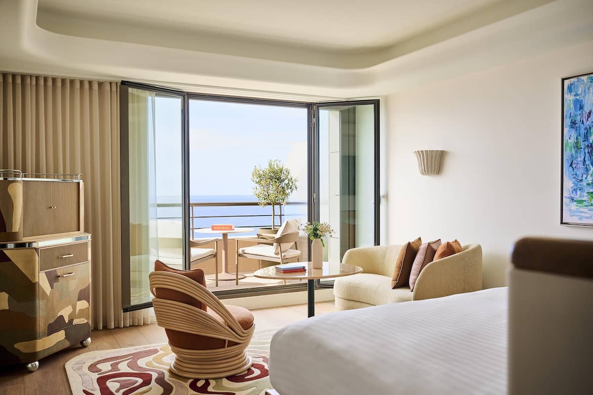 A hotel room with a bed, a sofa, a dining table, a chair, a lamp, a plant, and a view of the sea.