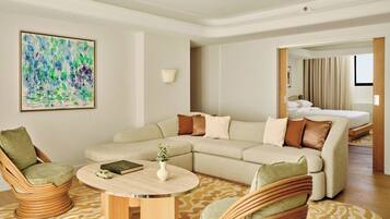 Quarto de hotel moderno com sofá modular, duas poltronas, mesa de centro redonda e cama com cortinas.