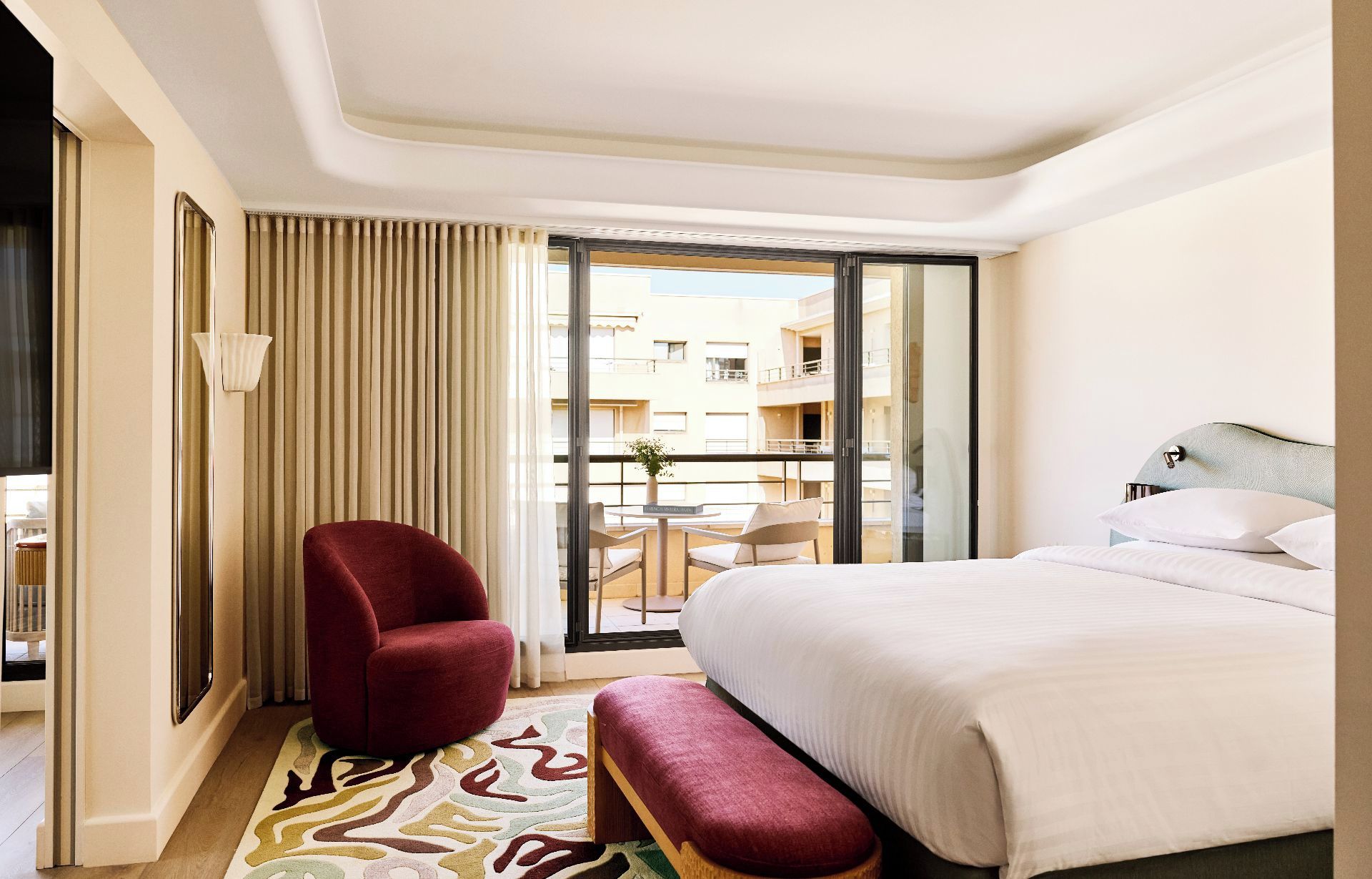 Quarto de hotel com uma cama, uma poltrona vermelha, um banco, uma mesinha e vista para a varanda.