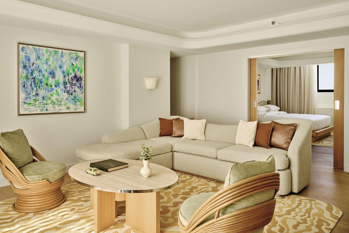 A modern hotel room with a sectional sofa, two wicker chairs, a round coffee table, a painting on the wall, and a bed in the background.