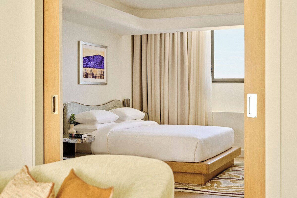 A hotel room with a bed, a bedside table with a lamp, a framed picture on the wall, and a window with curtains.