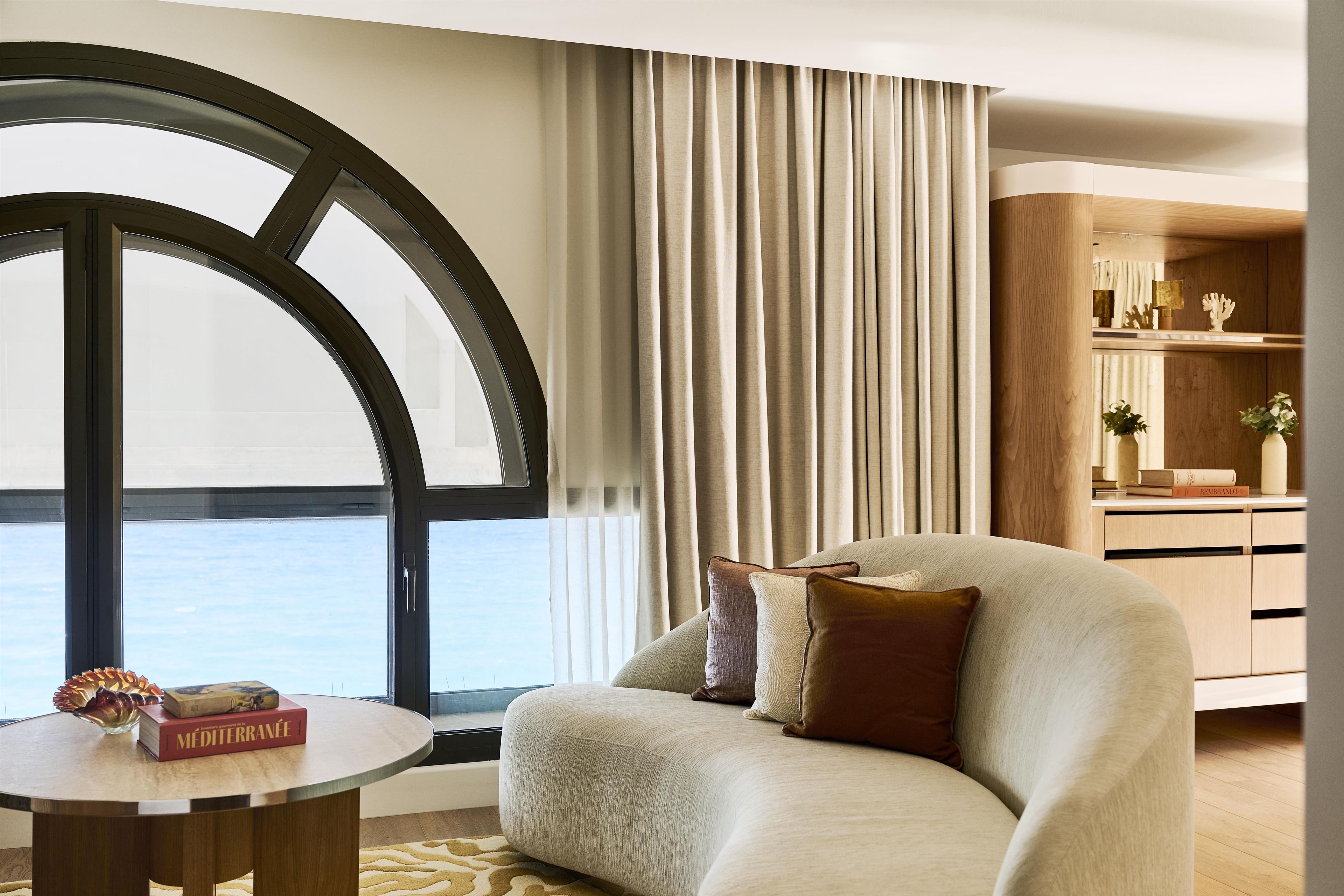 A modern living room with a large window, a sofa, a round table, and a bookshelf.