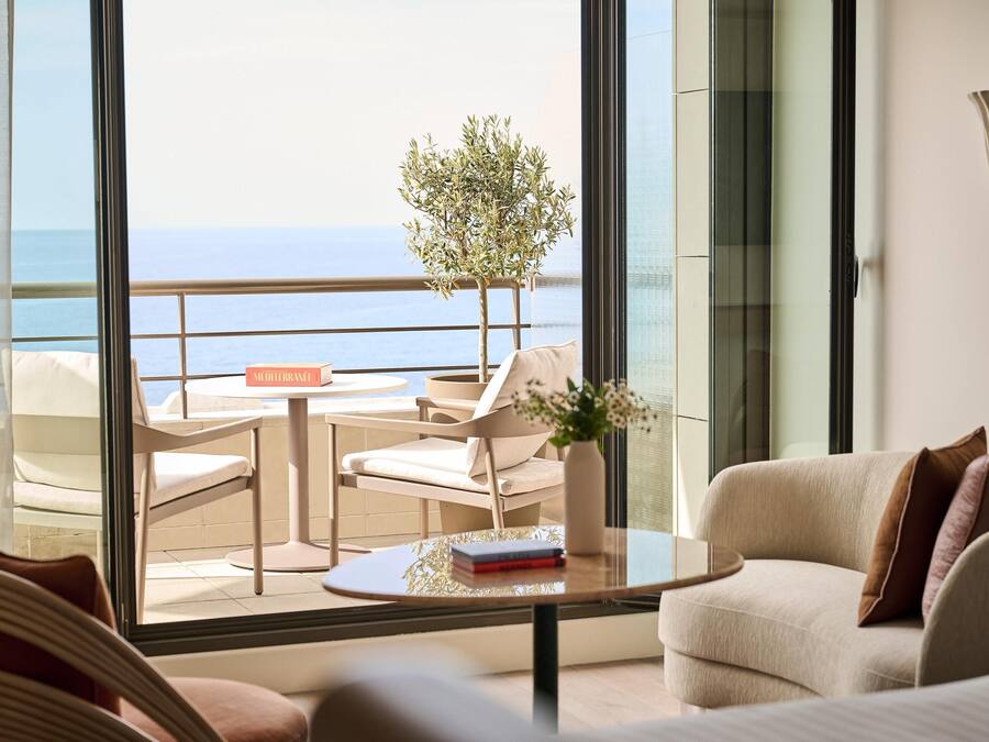 Quarto de hotel moderno com varanda, sofá, mesa com um livro e um vaso de planta.