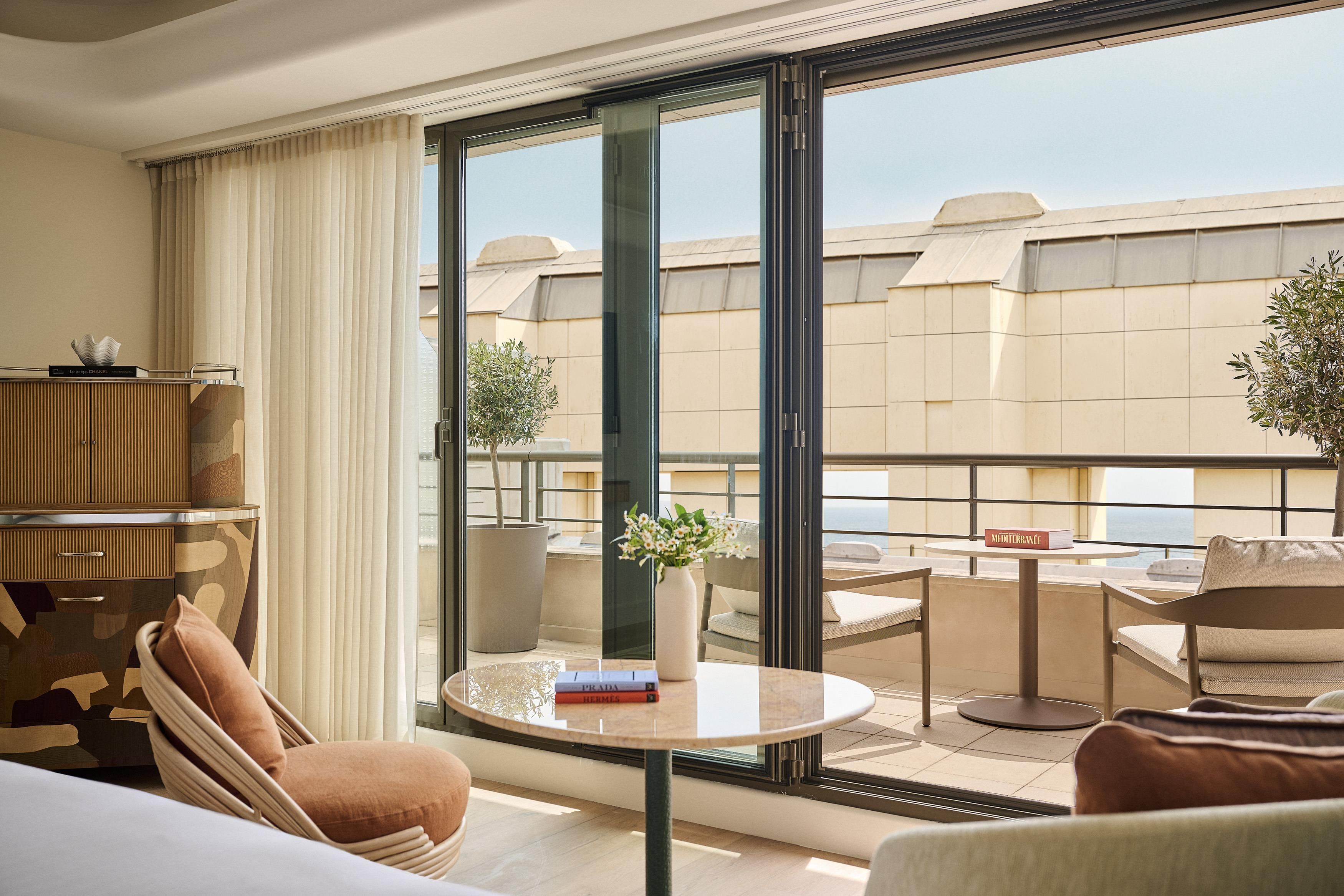 A hotel room with a balcony, a dining table, a sofa, a chair, a potted plant, and a minibar.