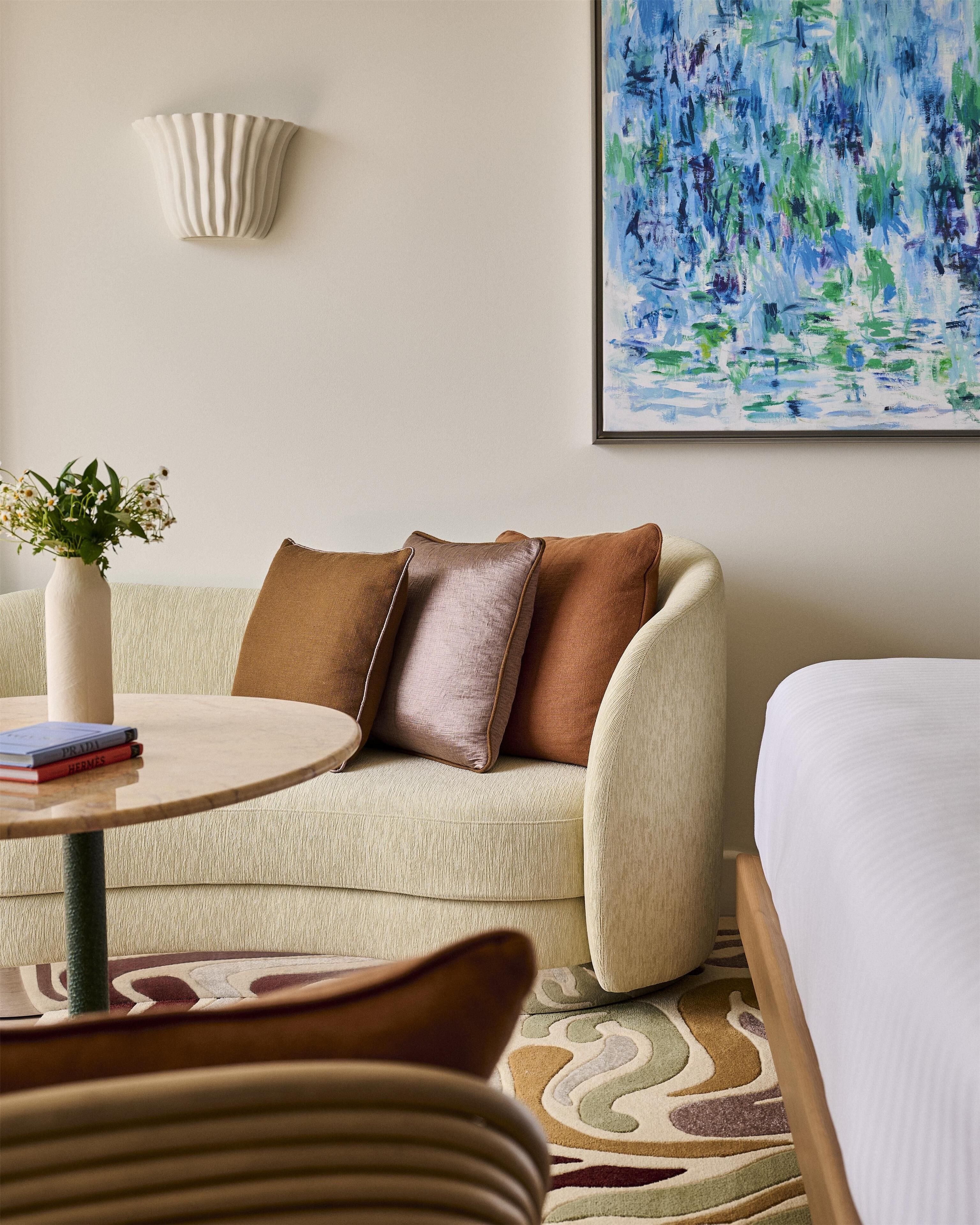 A hotel room with a sofa, a small round table with books, a vase with flowers, a painting on the wall, and a bed with white bedding.