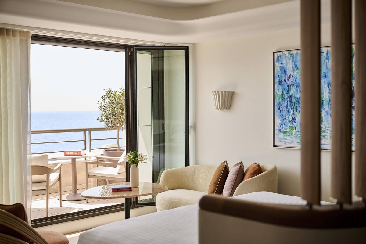 A hotel room with a balcony, a sofa, a dining table, and a painting.