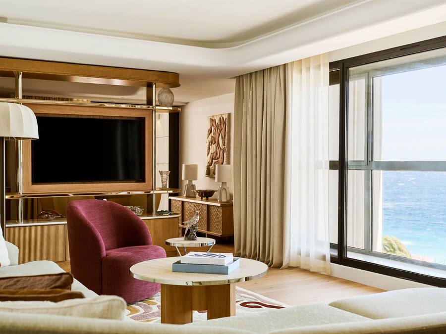 A modern living room with a large TV, comfortable seating, a small table, and a view of the sea.