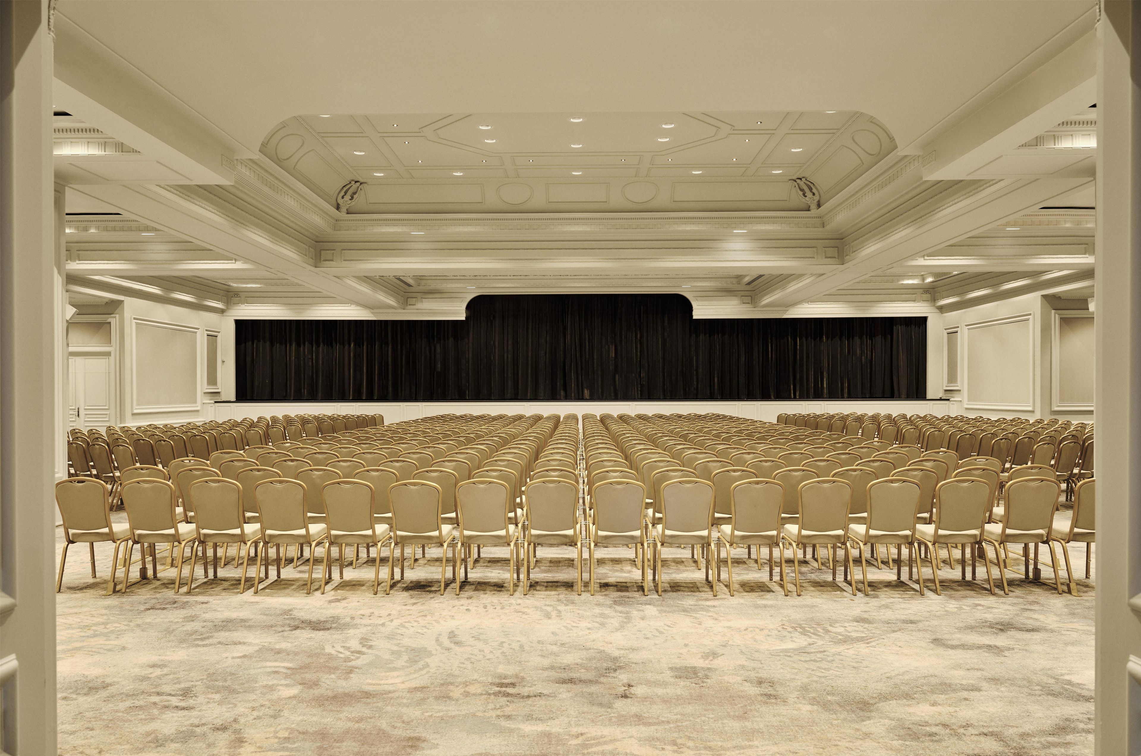 Un salón de conferencias con filas de sillas, un escenario con fondo negro e iluminación empotrada en el techo.