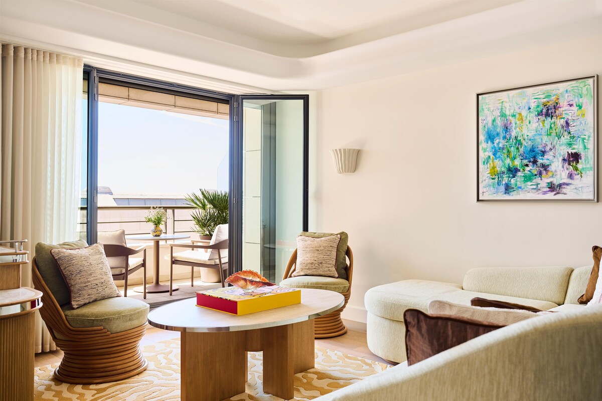 A modern living room with a round coffee table, two armchairs, a sofa, a dining table with chairs, a large window, and a colorful abstract painting on the wall.