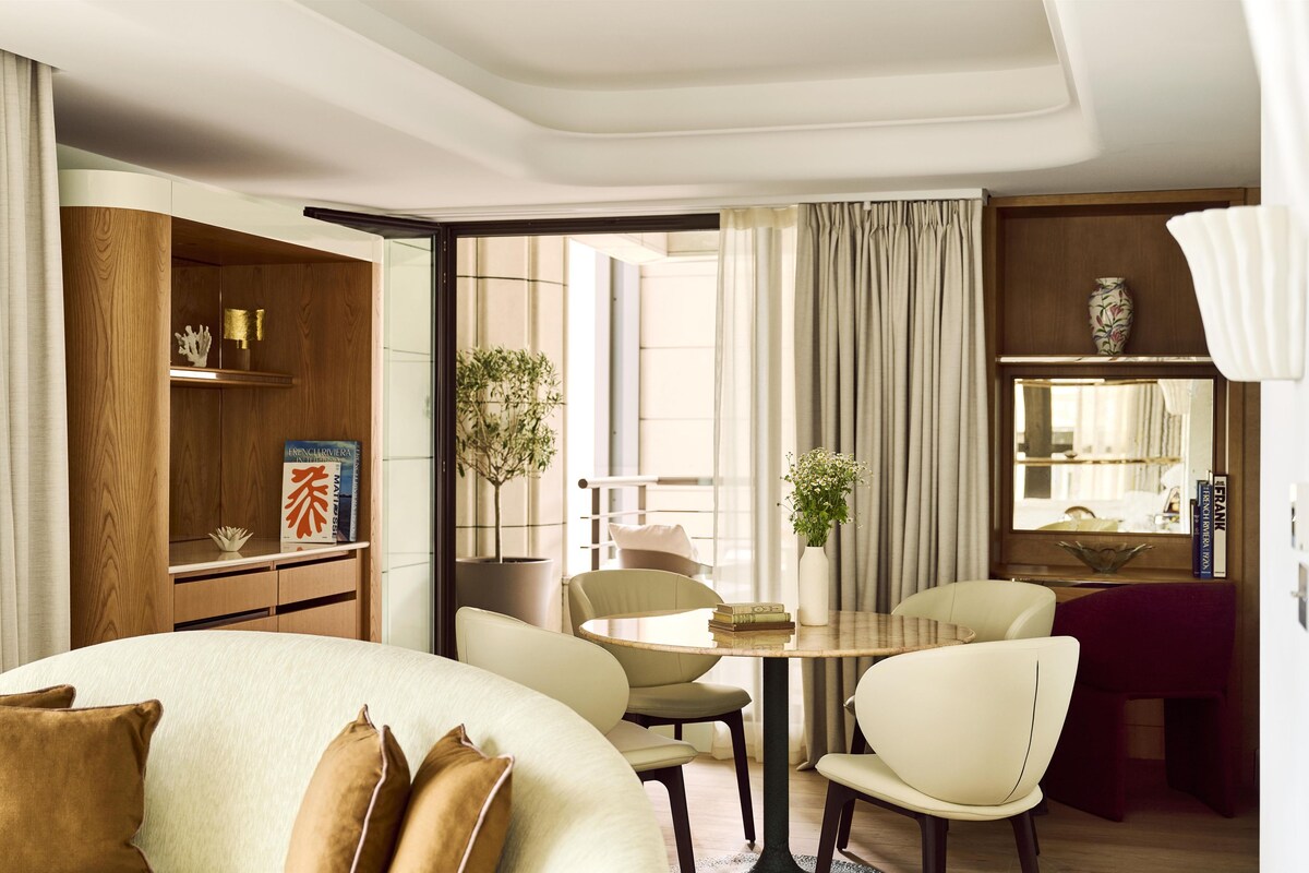 A hotel room with a bed, a dining table with chairs, a bookshelf, and a large window with curtains.