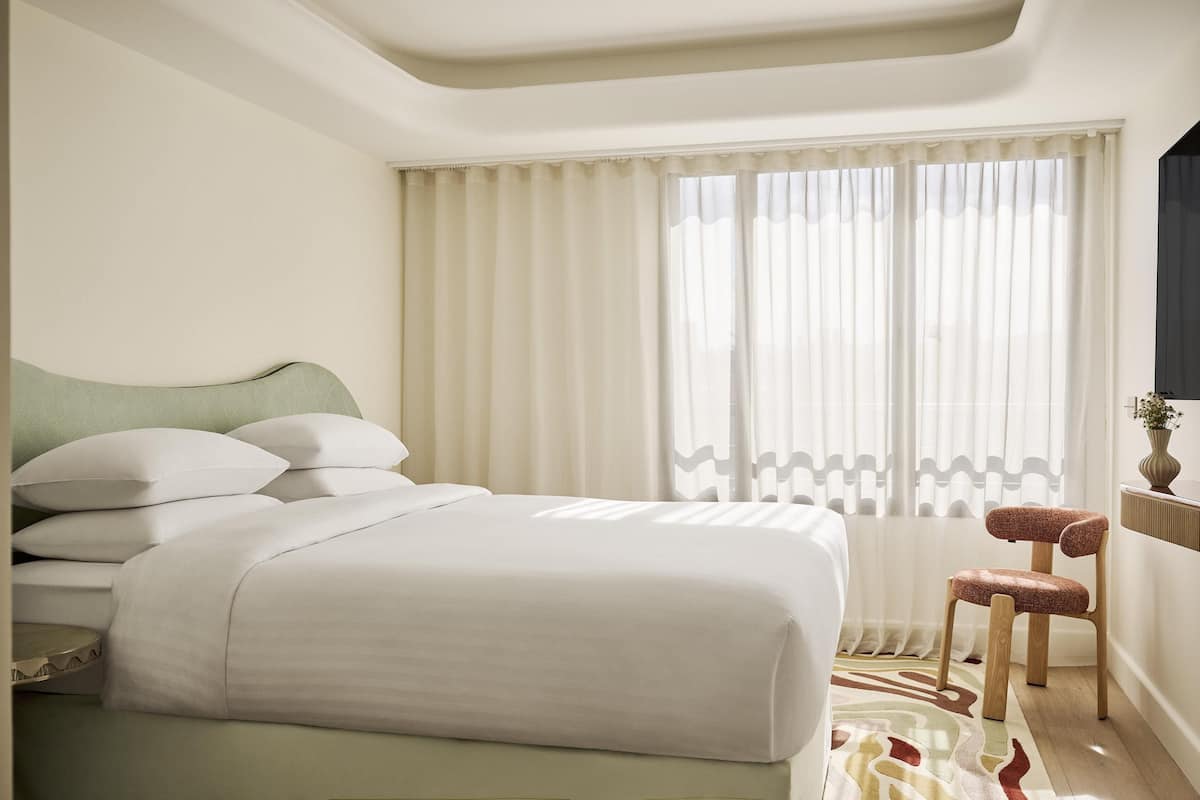 A hotel room with a bed, a chair, a window with curtains, and a wall-mounted TV.