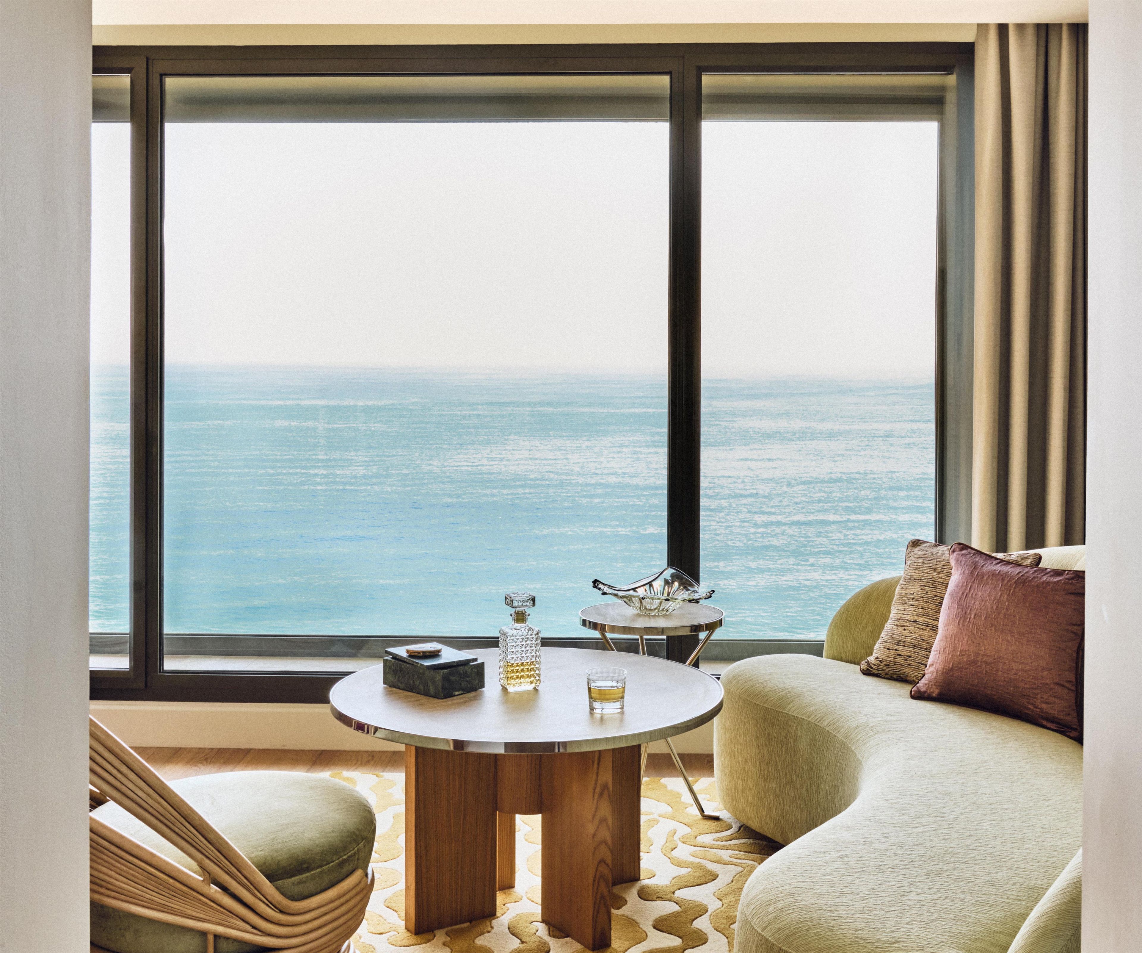 Una habitación con un ventanal grande que da al mar, una mesa redonda de madera con una botella y un vaso, y un sofá verde con un cojín.