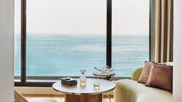Una habitación con un ventanal grande que da al mar, una mesa redonda de madera con una botella y un vaso, y un sofá verde con un cojín.