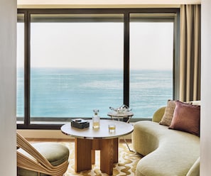 A room with a large window overlooking the sea, a round wooden table with a bottle and glass, and a green sofa with a cushion. - Hôtel Palais de la Méditerranée In The Unbound Collection By Hyatt (Nice)