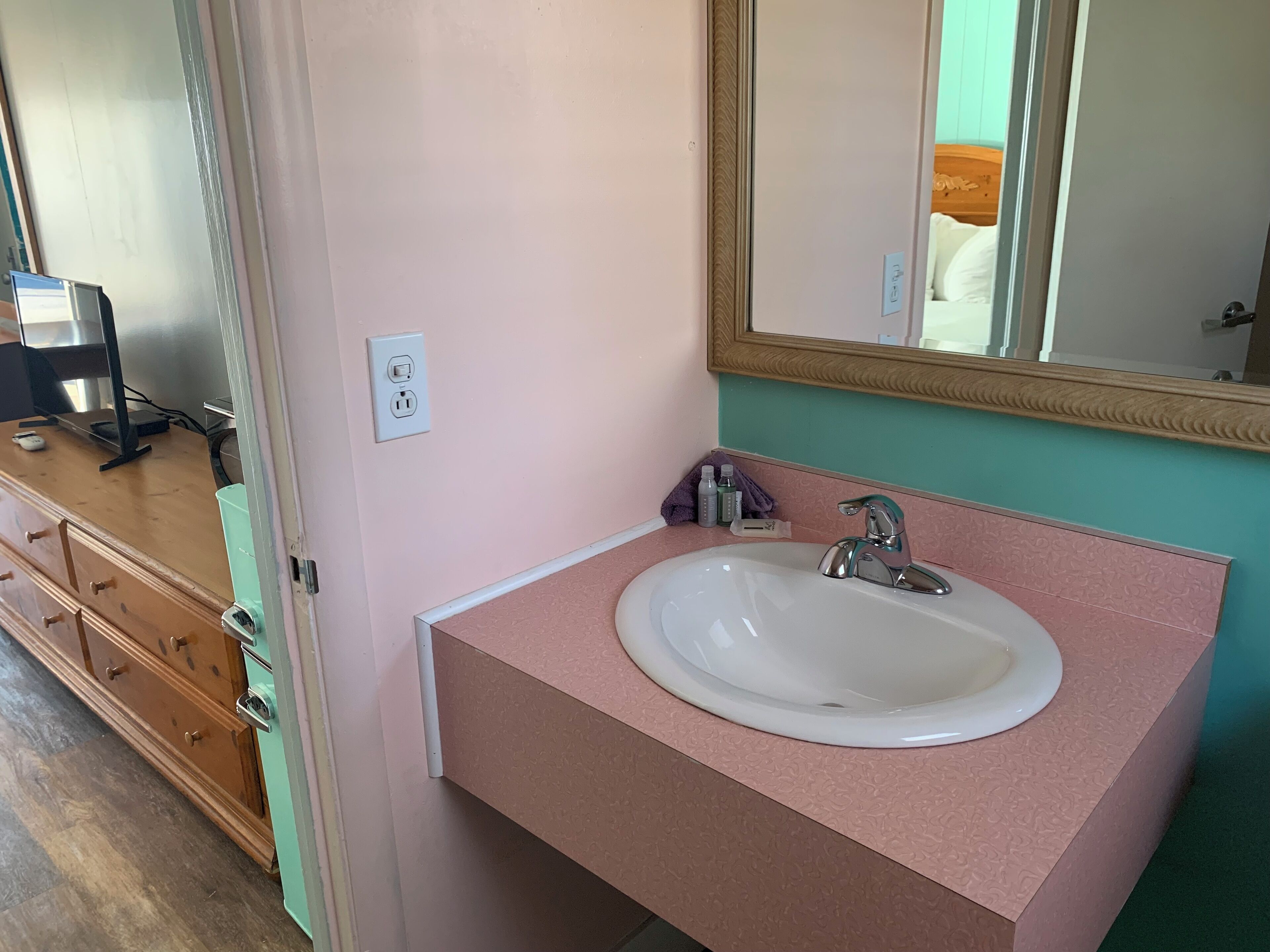Casa de banho com bancada rosa, lavatório branco e espelho. Tem uma cómoda de madeira com gavetas e uma pequena mesa com monitor de computador.