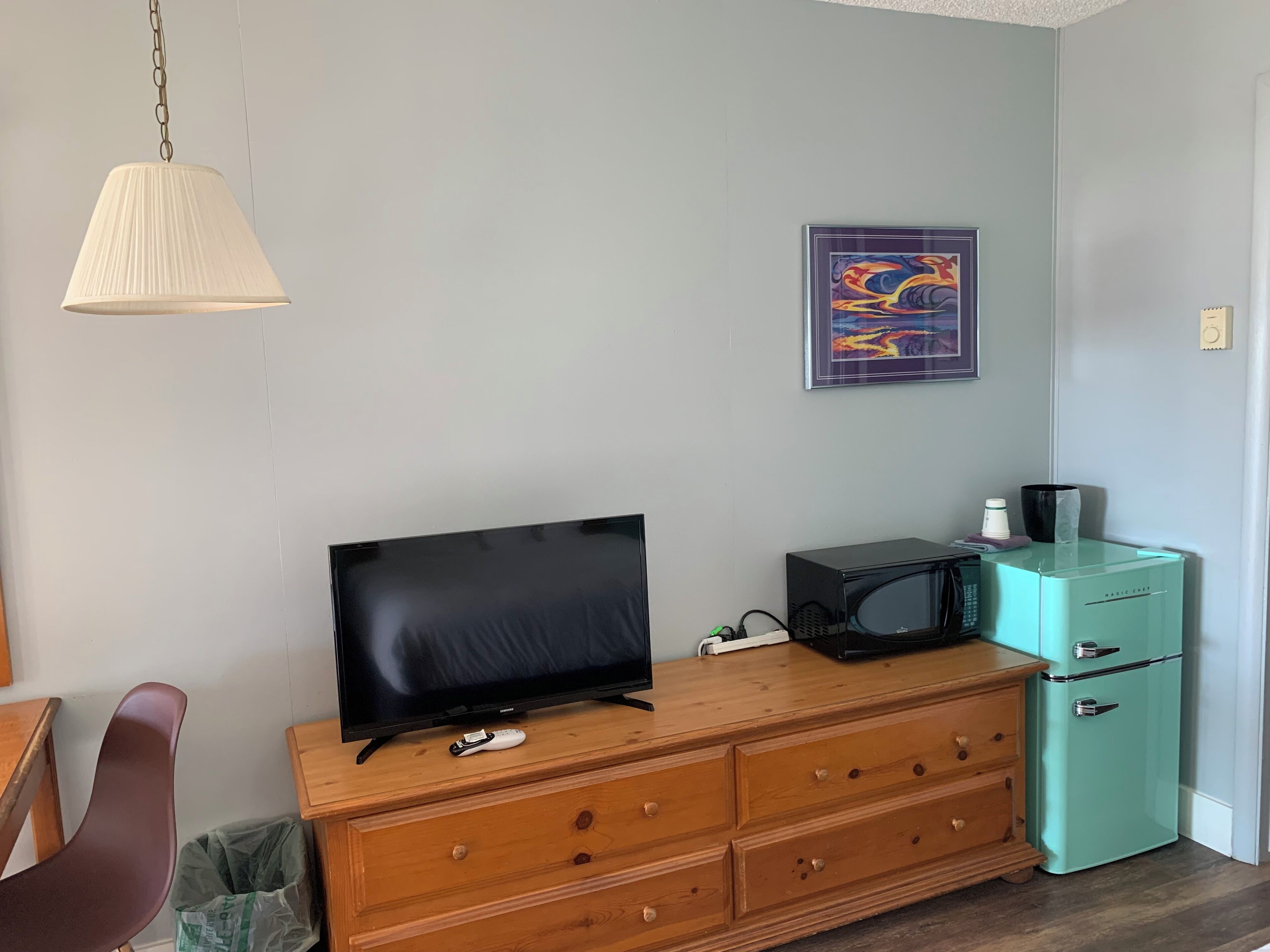 Una habitación con una televisión sobre un aparador de madera, un microondas, un cubo de basura y un refrigerador color aguamarina.
