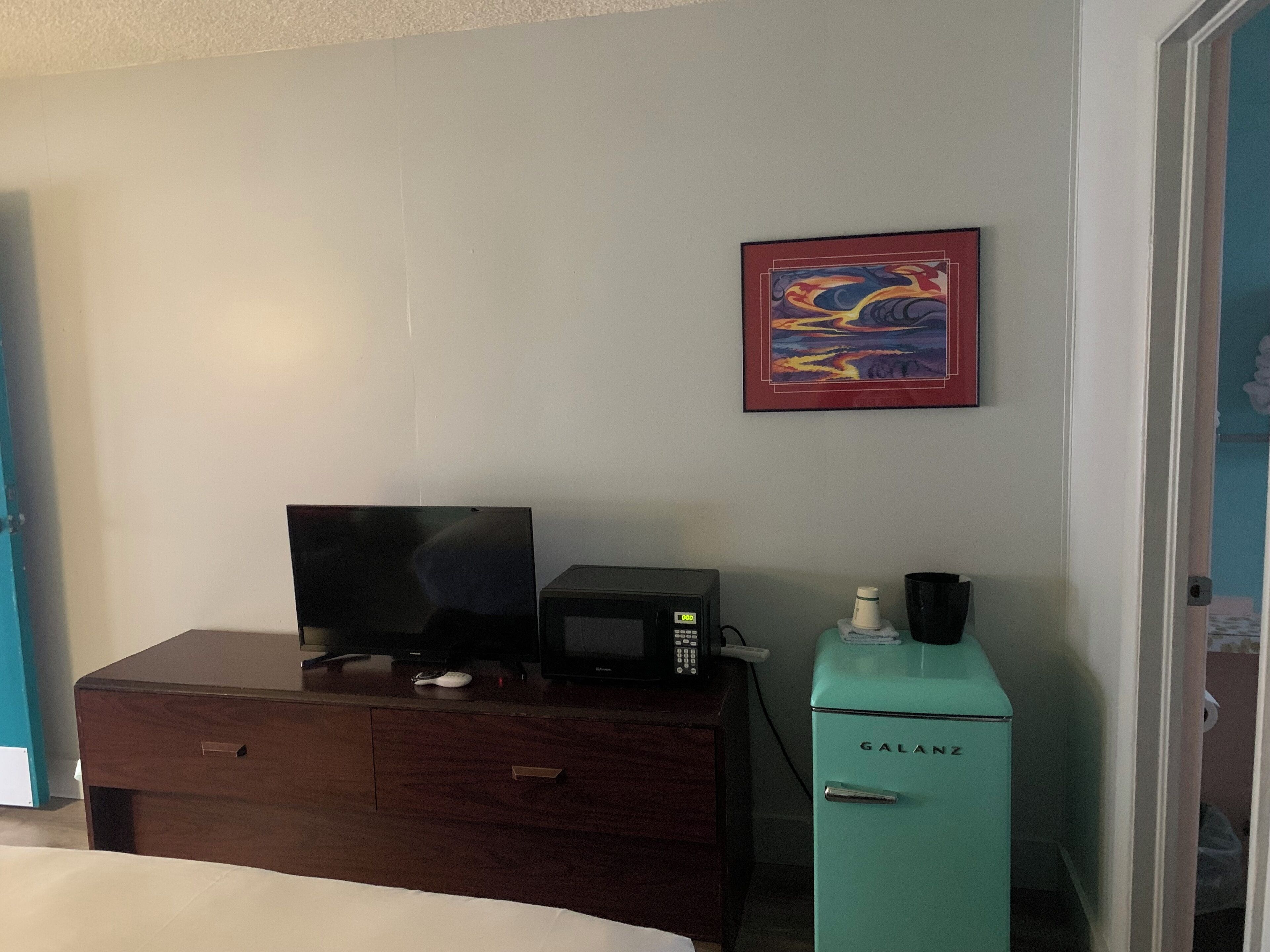 A hotel room with a bed, a desk with a computer, a microwave, and a teal-colored refrigerator.