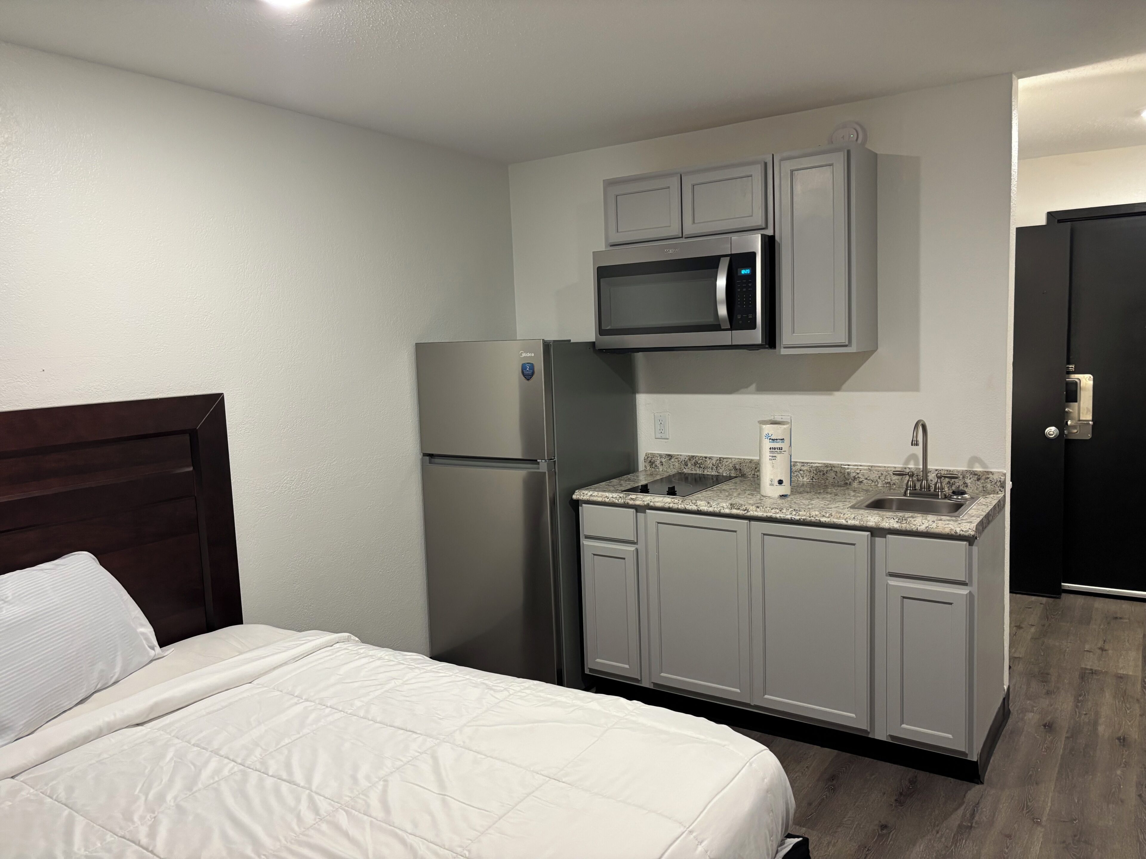 A compact hotel room with a bed, a kitchenette including a refrigerator, microwave, and sink, and a dark door.
