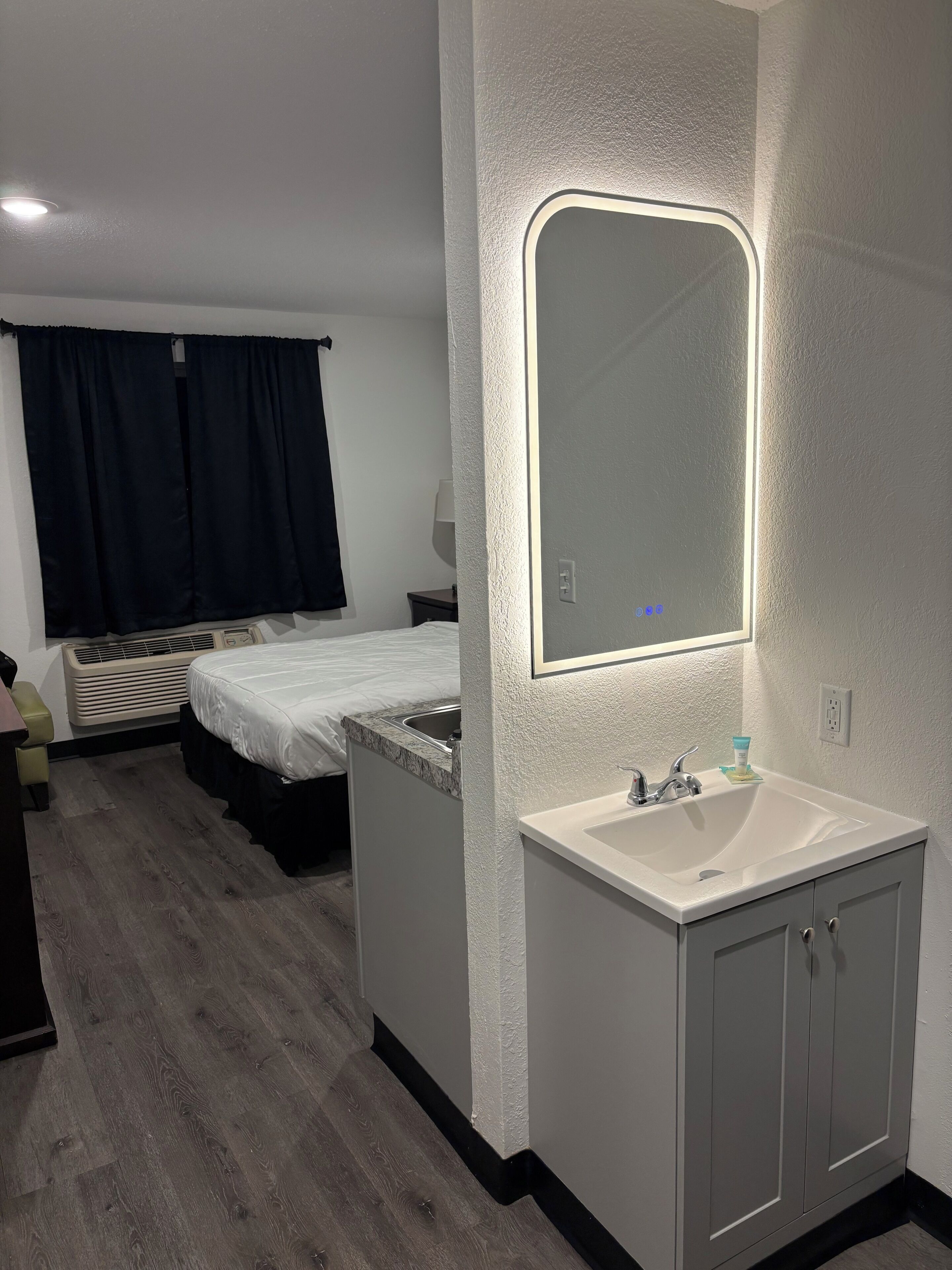 Une chambre d’hôtel avec un lit, une table de chevet, un miroir, un lavabo et une fenêtre avec des rideaux.