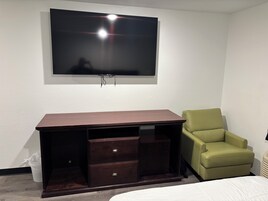Una habitación con una televisión de pantalla plana montada en la pared, un escritorio de madera con cajones, una silla verde y una cama con ropa blanca.