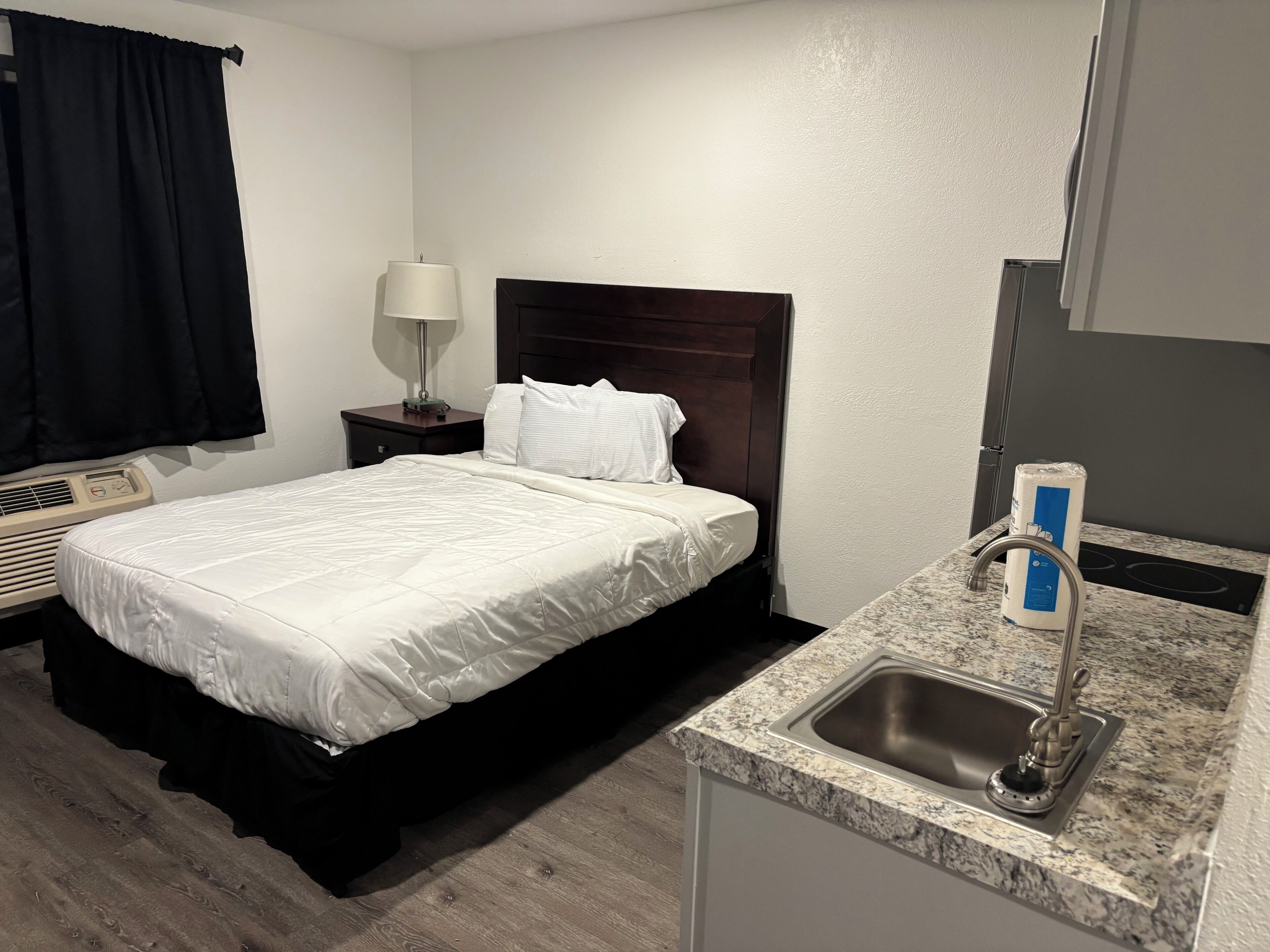 Habitación de hotel con cama, mesita de noche con lámpara, unidad de aire acondicionado integrada, kitchenette pequeña con fregadero y ventana con cortinas negras.