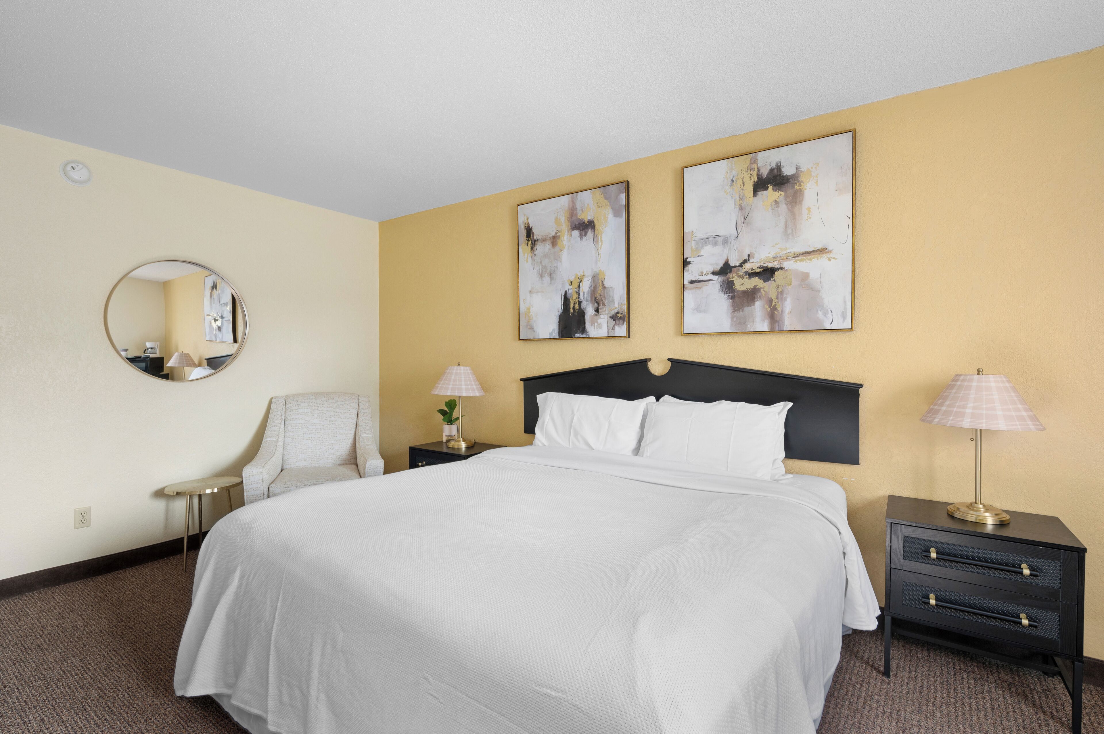 A hotel room with a large bed, a round mirror, two bedside tables with lamps, and abstract wall art.