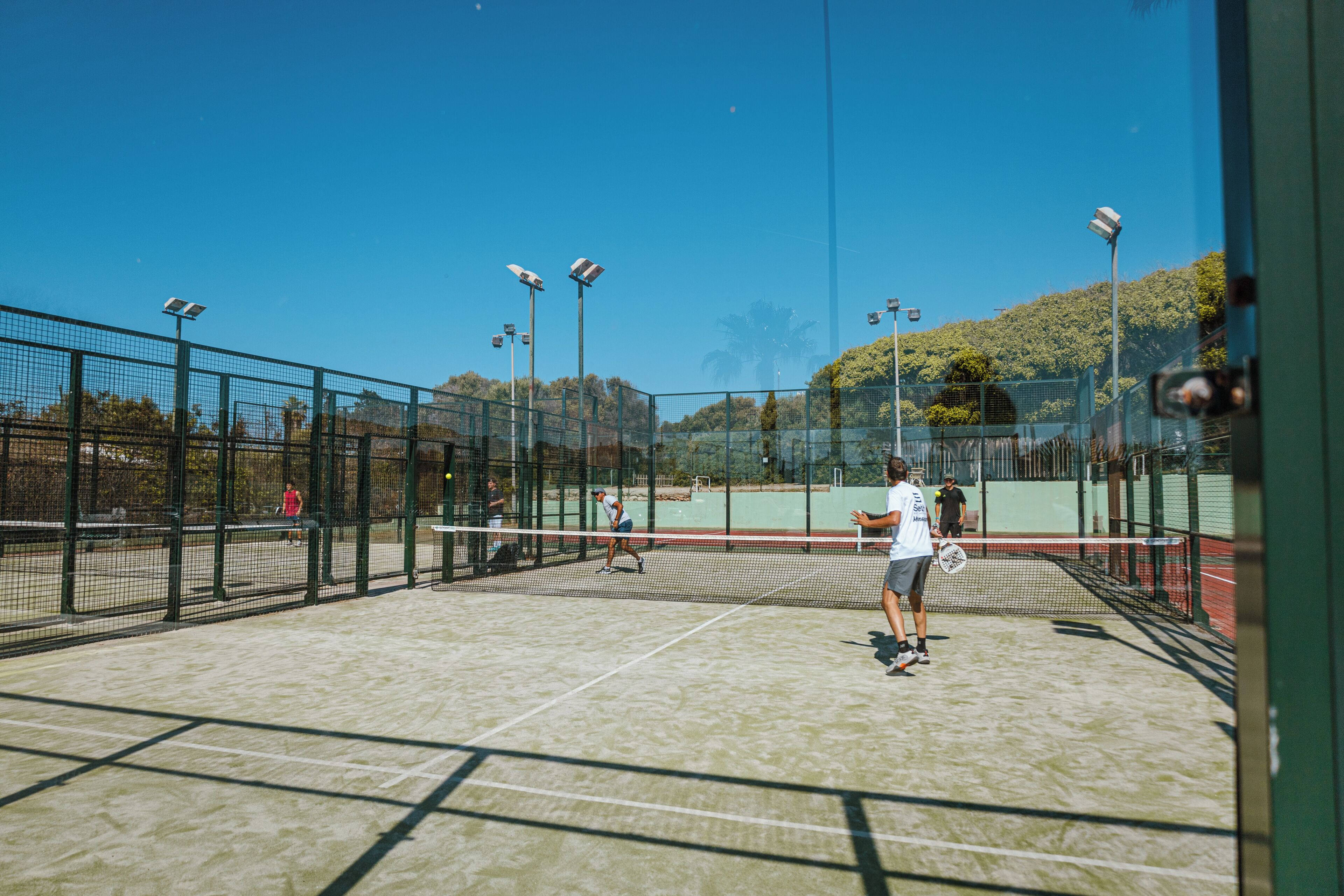 Una cancha de tenis con red, rodeada por una valla, y equipada con iluminación artificial.