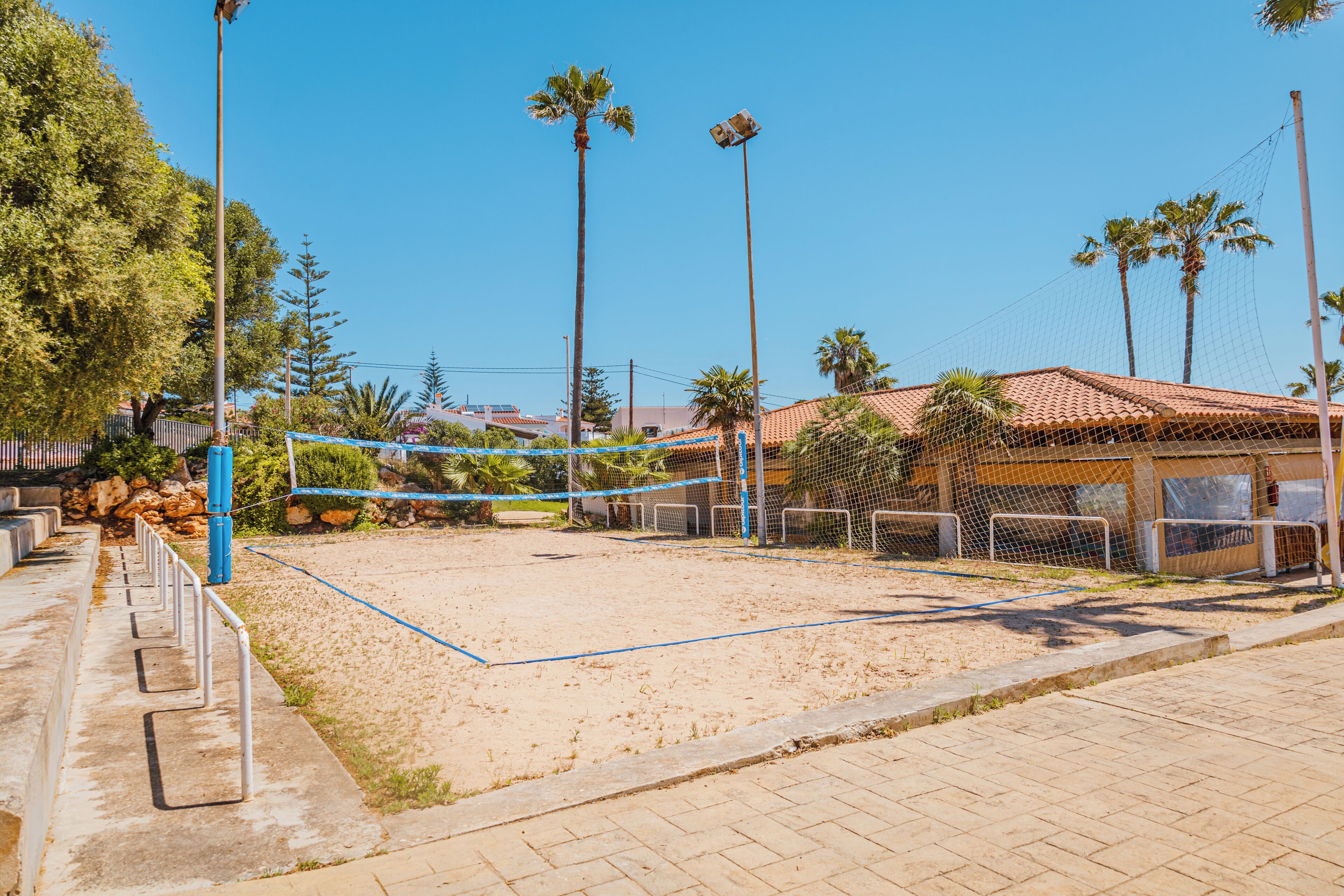 A sandy volleyball court with a net, surrounded by palm trees and a building with a tiled roof.