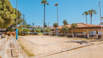 A sandy volleyball court with a net, surrounded by palm trees and a building with a tiled roof.