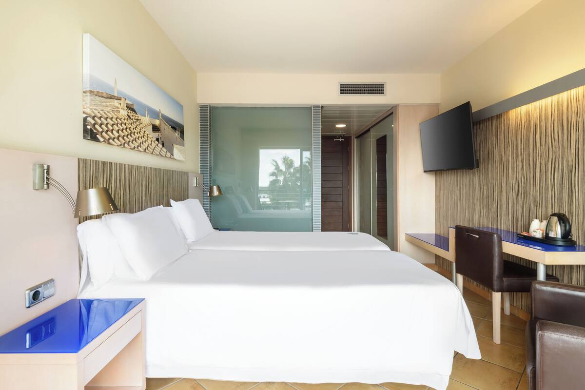 Une chambre d’hôtel moderne dotée d’un grand lit, d’un bureau avec une chaise, d’un téléviseur à écran plat fixé au mur et d’une grande fenêtre donnant sur des palmiers.