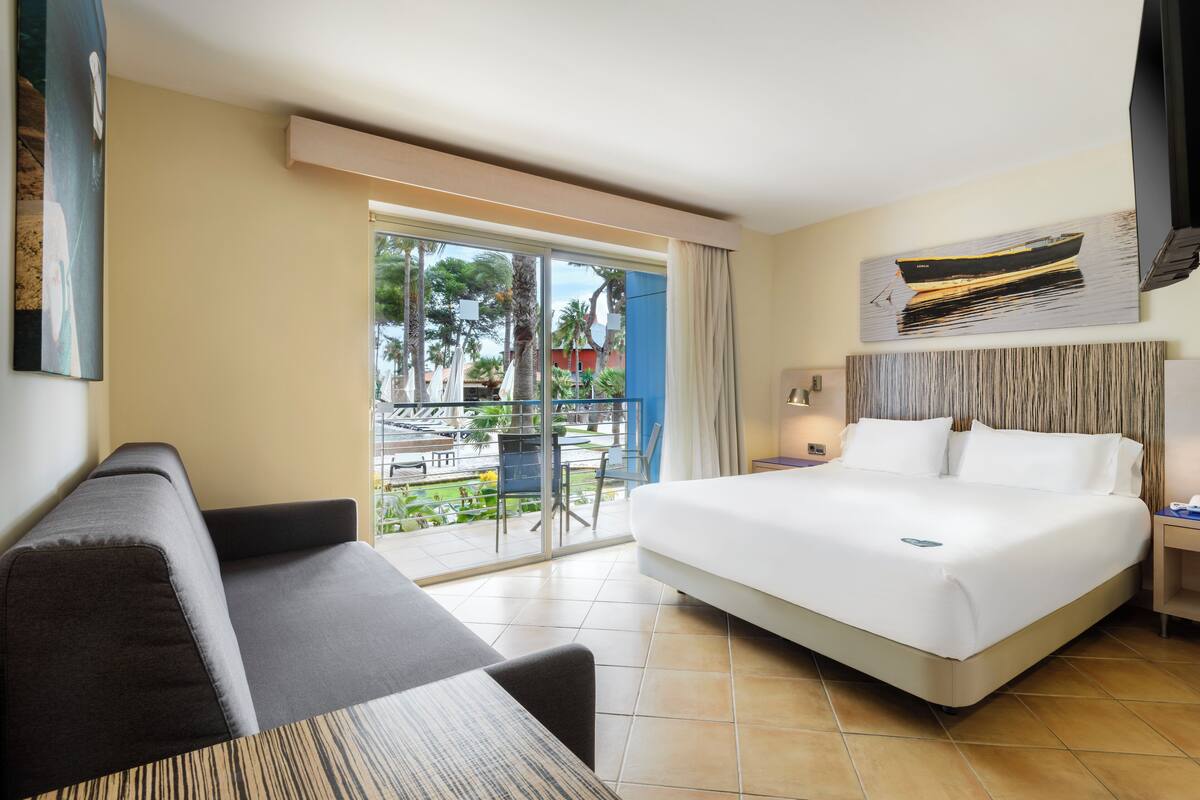 Une chambre d’hôtel avec un grand lit, un canapé, une table à manger et un balcon offrant une vue sur les palmiers et une piscine.