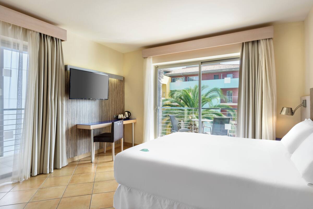 Une chambre d’hôtel avec un lit, un bureau, une télévision et un balcon offrant une vue sur les immeubles et les palmiers.