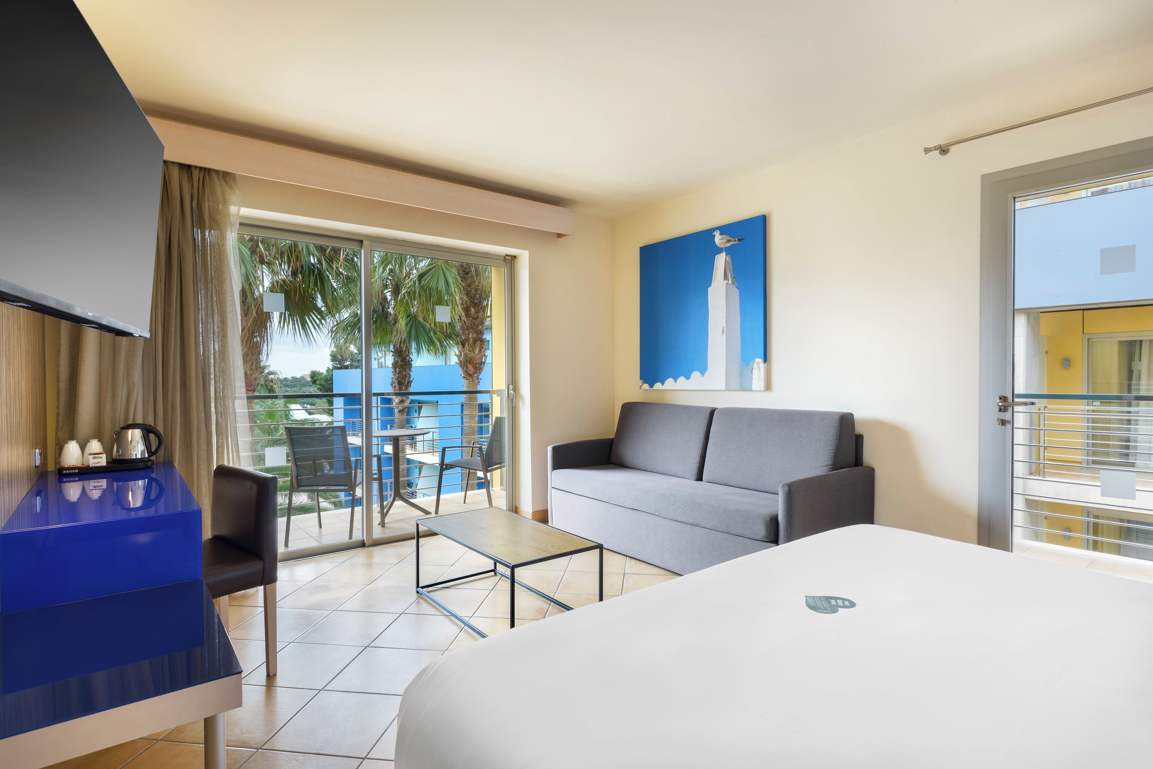 Habitación de hotel con cama, sofá, una mesita, televisor y balcón con sillas y una mesa.