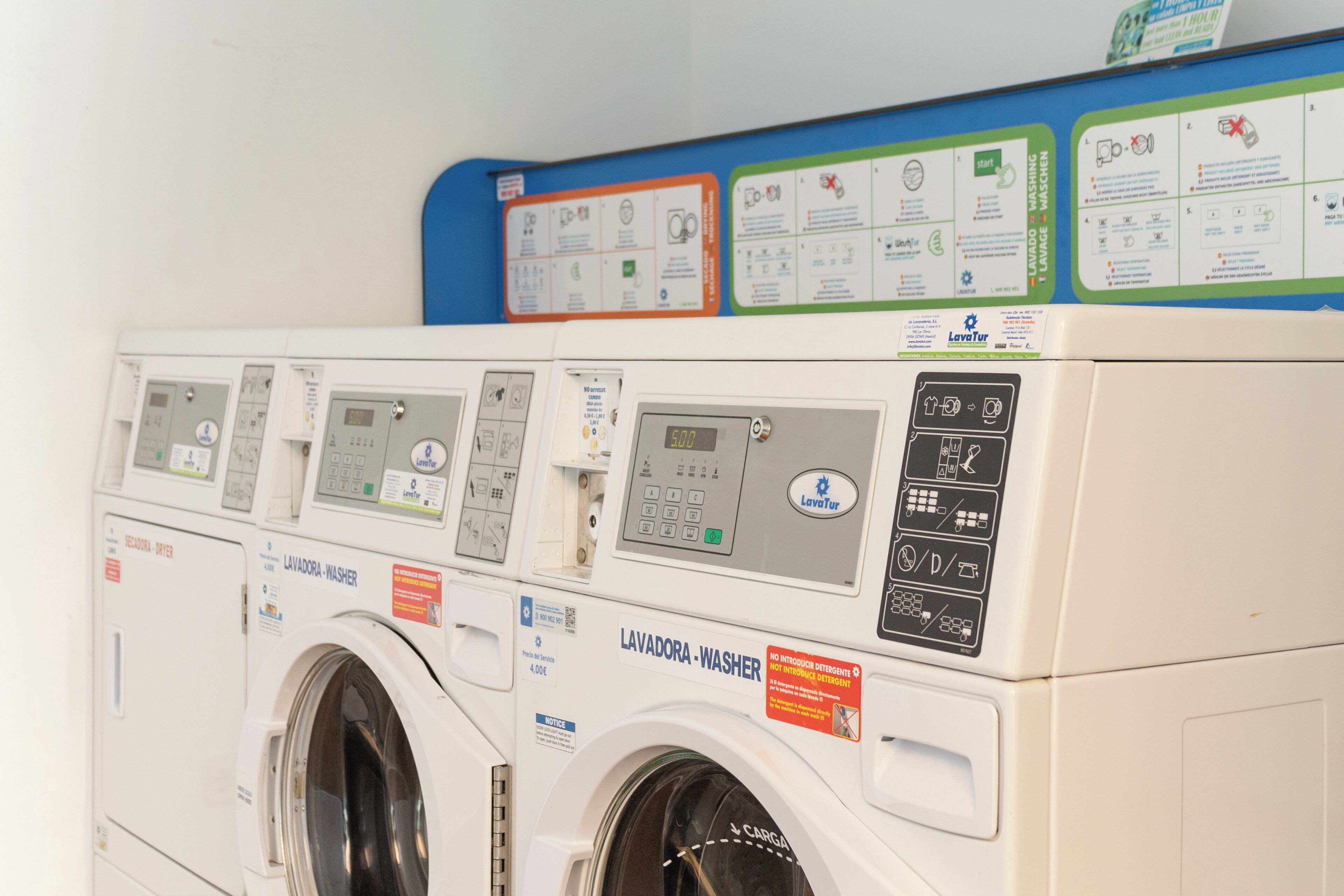 A row of white laundry machines with control panels and instructional signs on a wall.