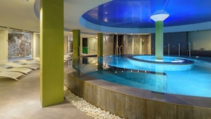 A modern indoor swimming pool with a glass enclosure, a lounge area with chairs, and a stone wall.
