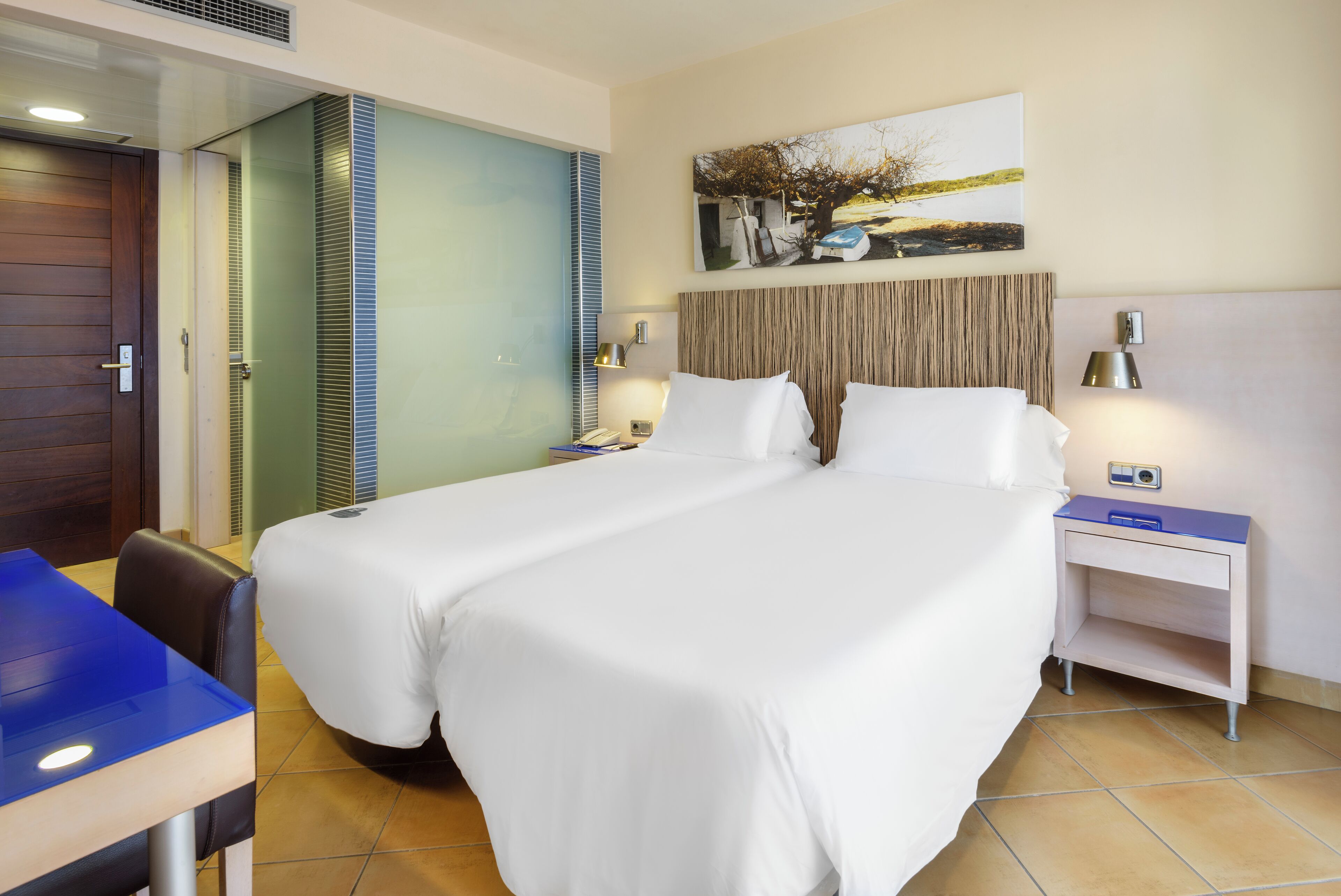 A hotel room with a large bed, a desk, a chair, a nightstand, and a painting on the wall.
