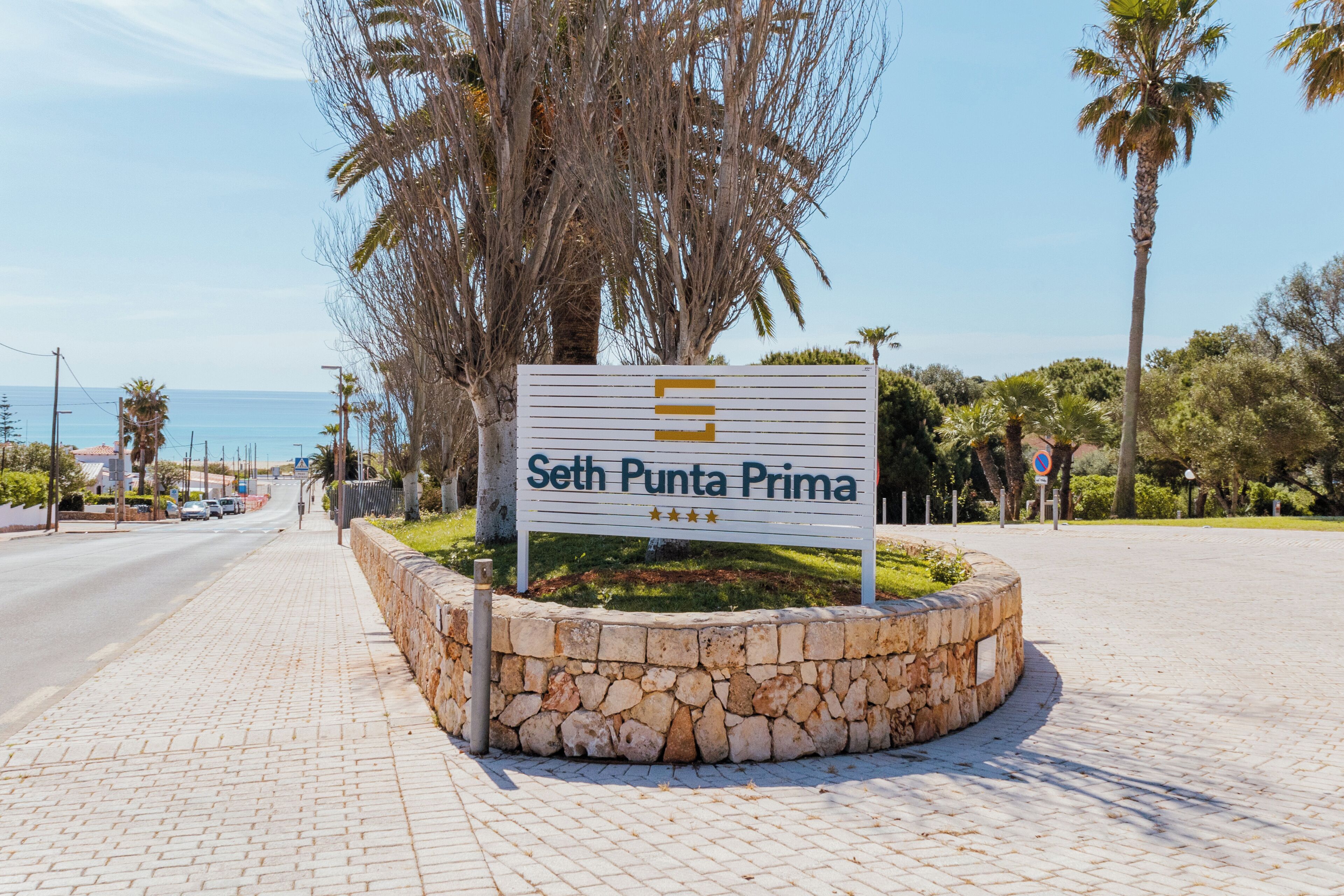 Señal de 'Seth Punta Prima' con borde de piedra, palmeras y cielo despejado.