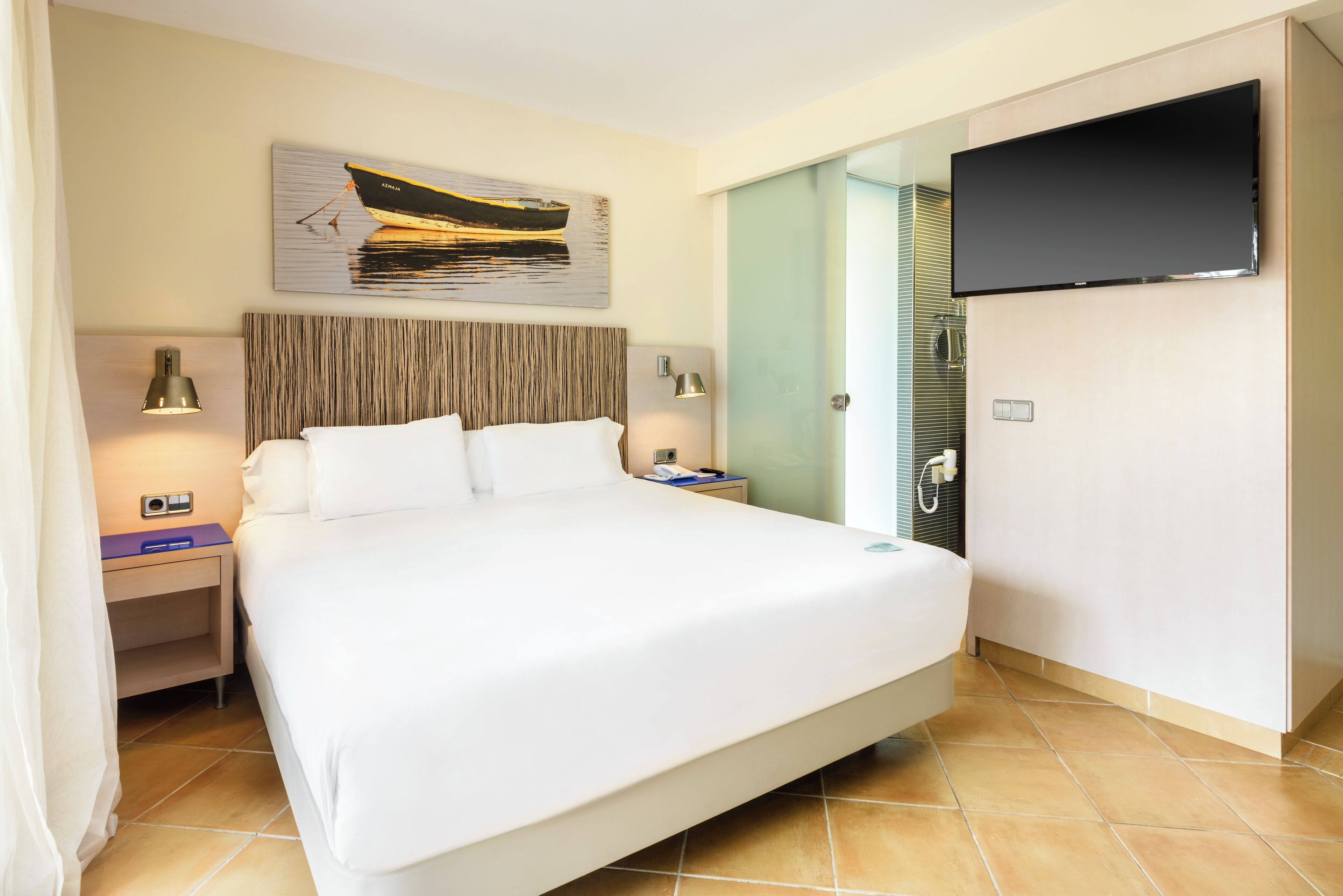 Ein Hotelzimmer mit einem großen Bett, einem an der Wand befestigten Flachbildfernseher, einem Bild eines Bootes an der Wand und einem Nachttisch mit Lampe und Telefon.