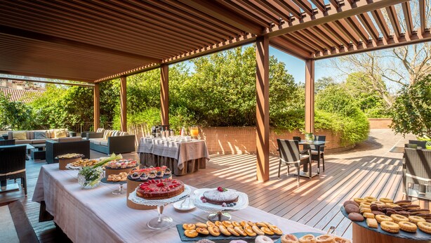 Un área de comedor al aire libre con una pérgola cubierta, una variedad de pasteles y postres en una mesa de buffet, y asientos exteriores con mesas y sillas.