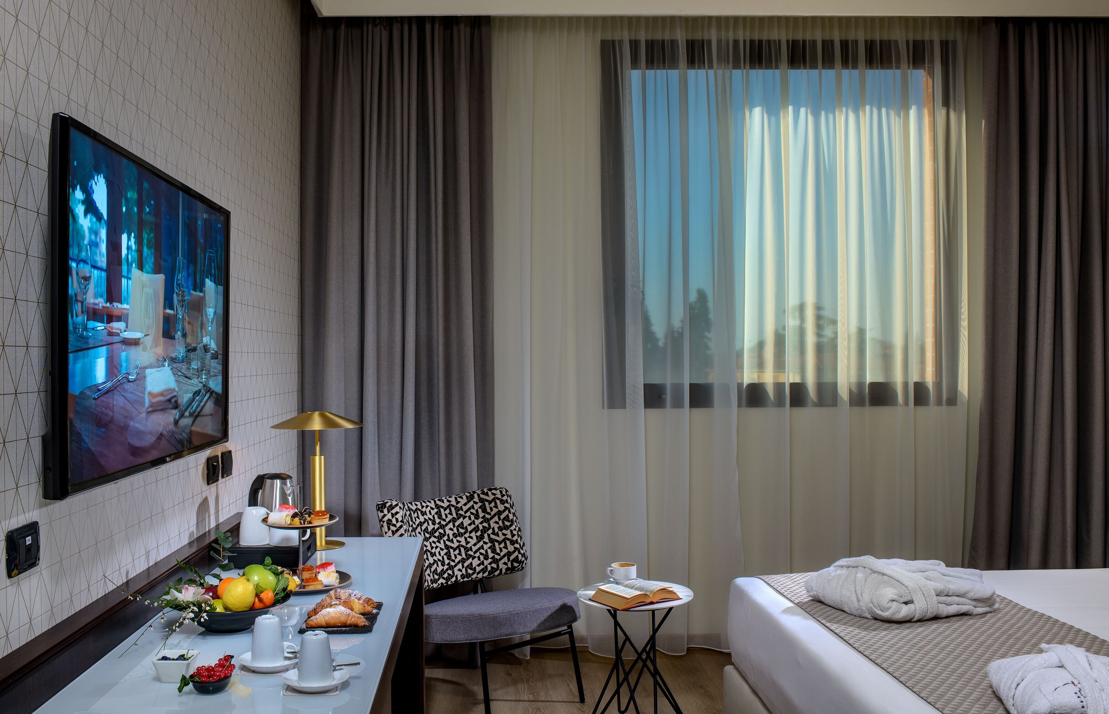 A hotel room with a bed, a desk with a laptop, a chair, a small table with a book and a cup, and a large window with curtains.