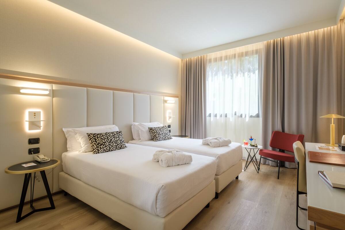 A hotel room with two beds, a desk, a chair, and a small table with a lamp.