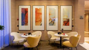 Eine moderne Hotelhalle mit vier gerahmten abstrakten Gemälden an der Wand, einer Sitzecke mit Korbstühlen und einem runden Tisch sowie einem Empfangsschalter.