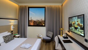 Habitación de hotel con cama, escritorio, silla, televisor y vista panorámica de la ciudad a través de una ventana.