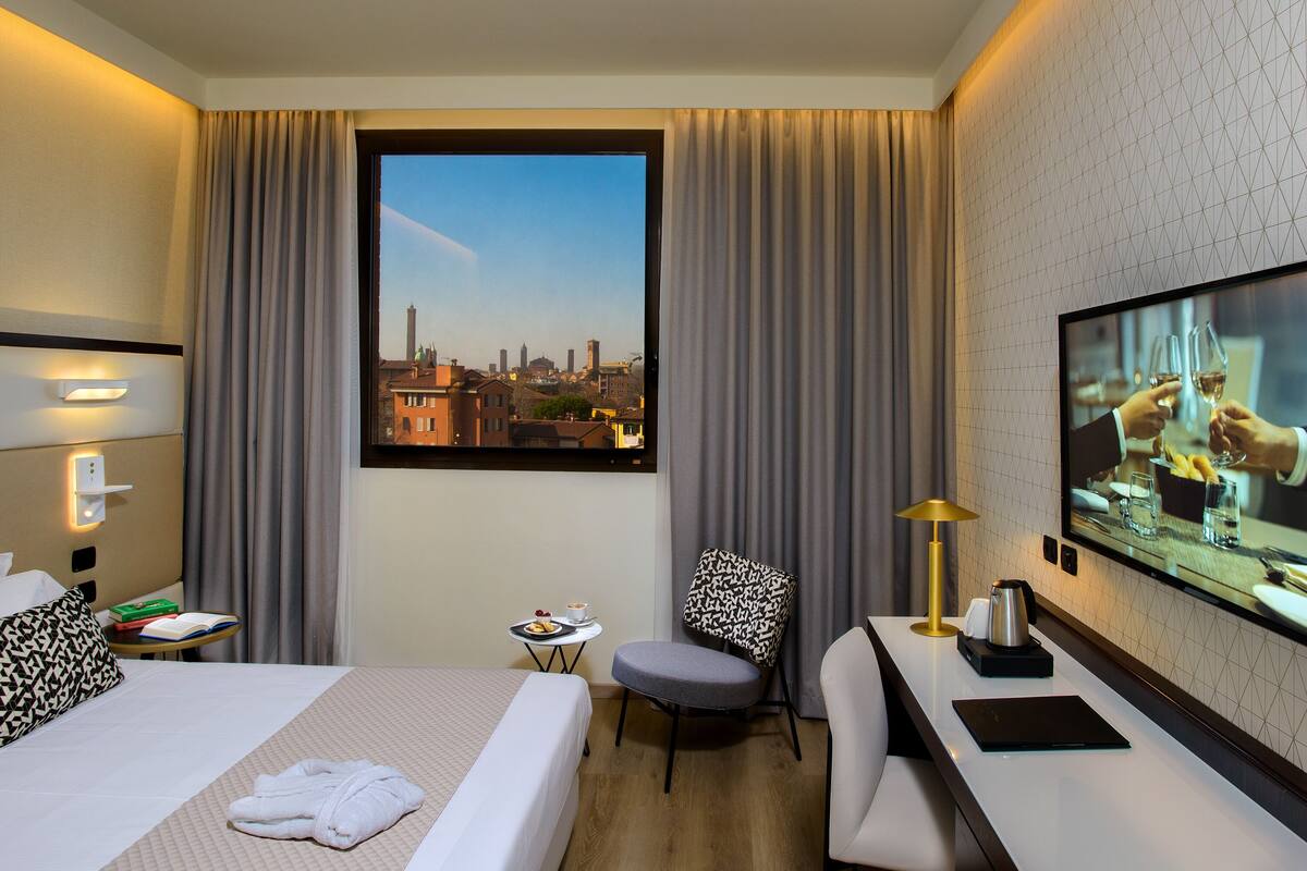 A hotel room with a bed, a desk, a chair, a TV, and a view of a cityscape through a window.